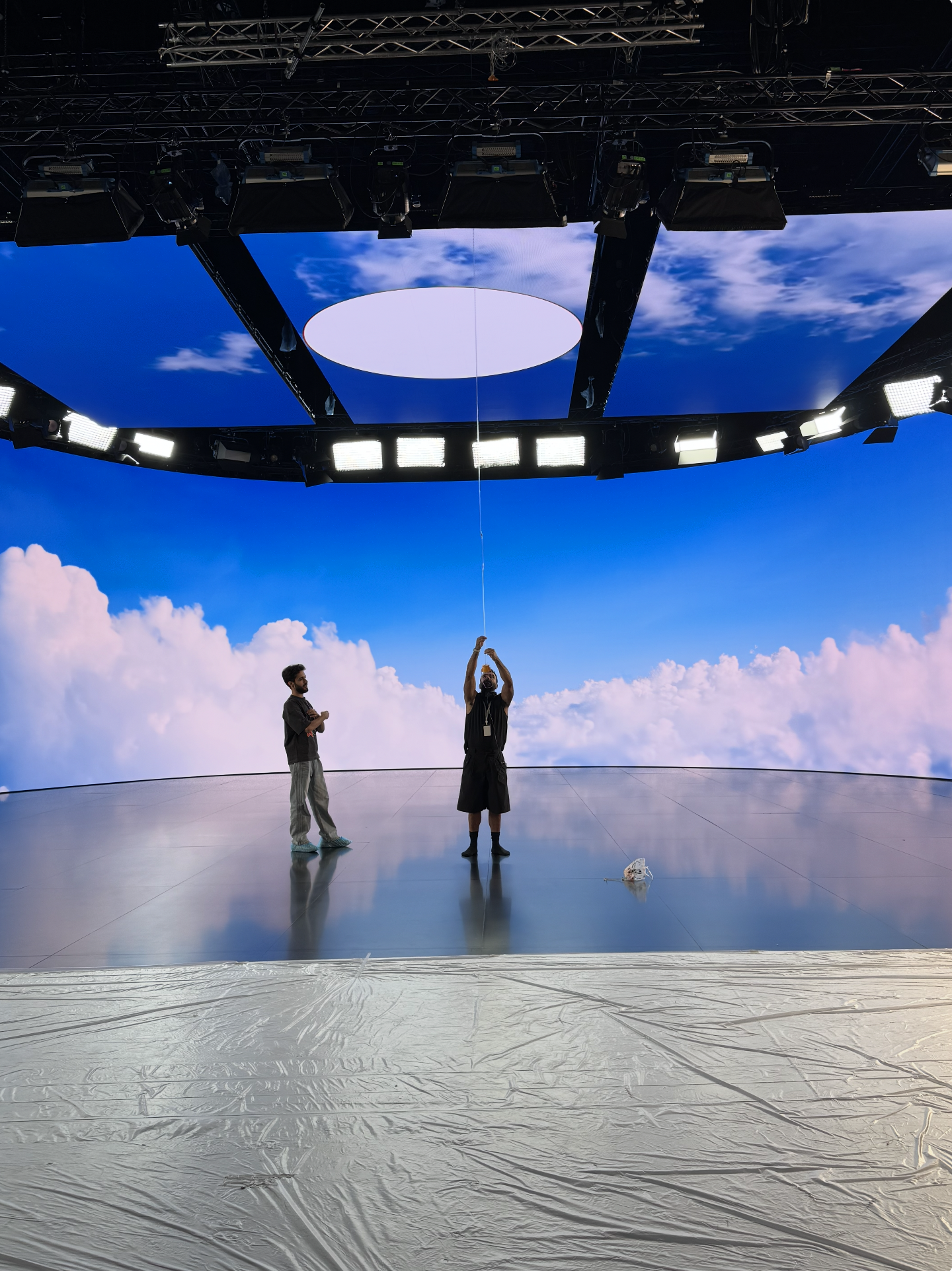 Deux hommes dans une salle de contrôle ou un studio, une personne tient un ballon ou un objet suspendu au plafond, avec un fond d'écran numérique affichant des nuages et un ciel bleu.
