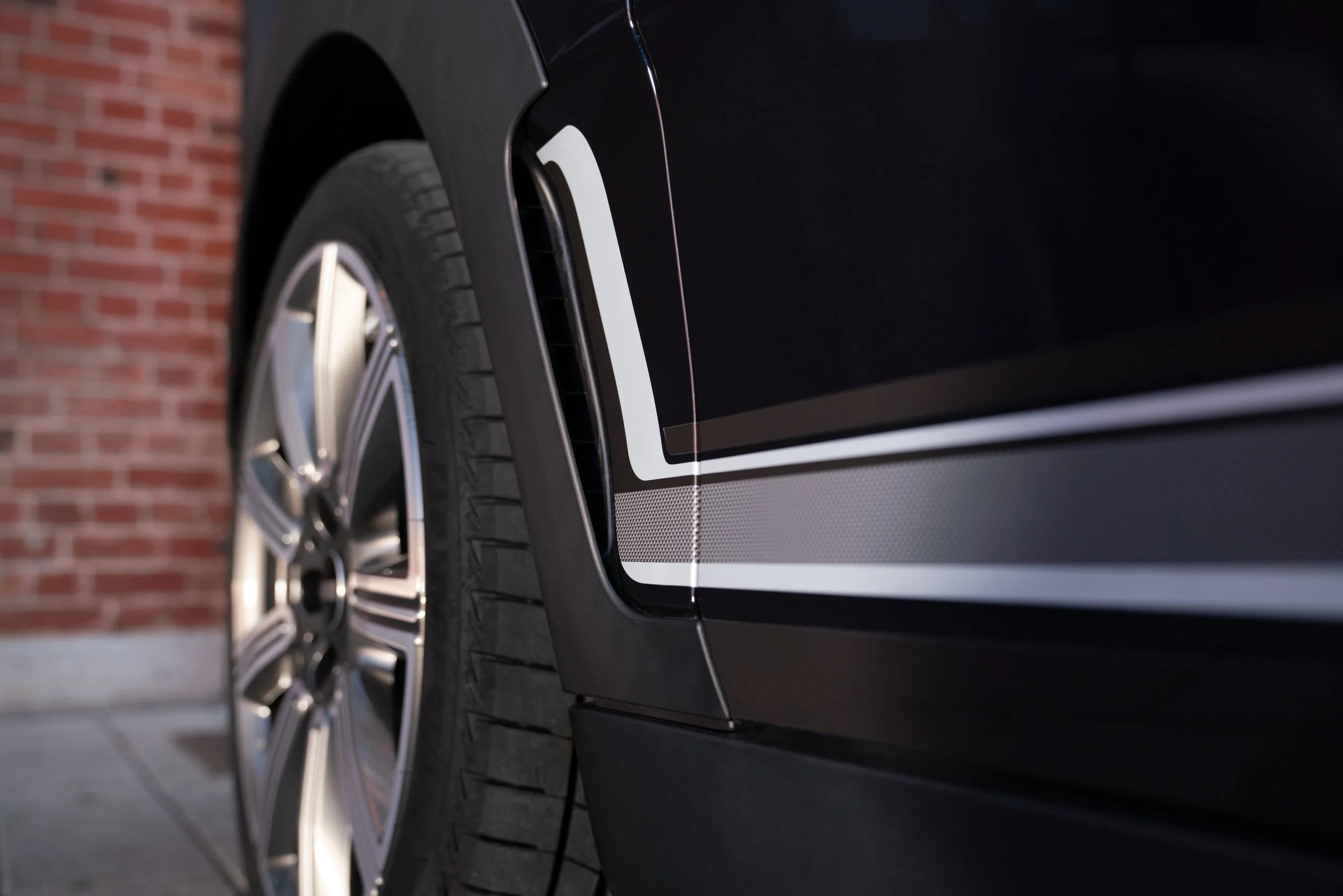 Détail d'une voiture noire, montrant la roue et la partie latérale, avec un mur de briques en arrière-plan.