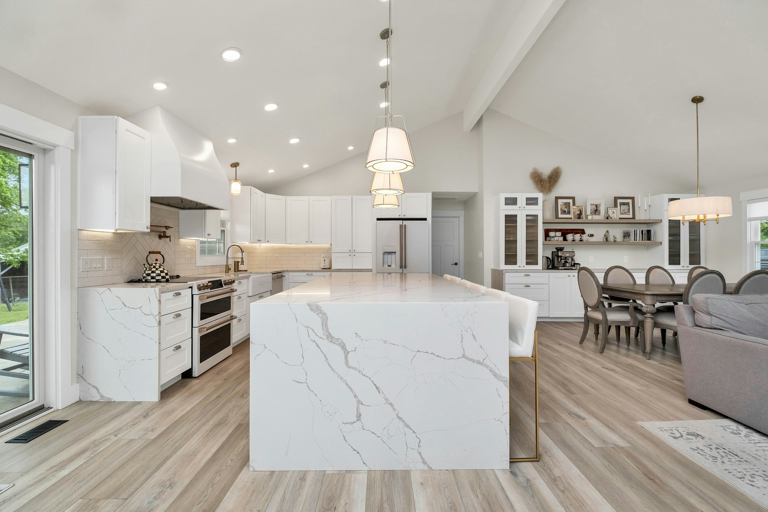 Bright, modern kitchen with white cabinets, marble island, and light wood flooring, open to dining and living areas.