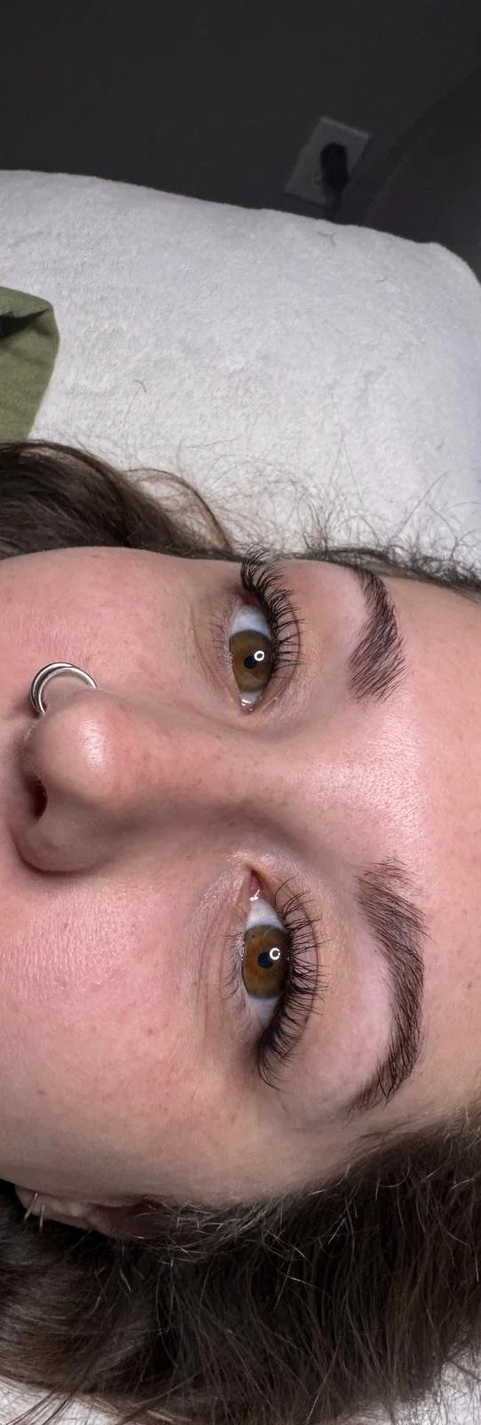 Close-up of a woman's face with brown eyes, dark eyebrows, and nose piercing, lying on a white surface.
