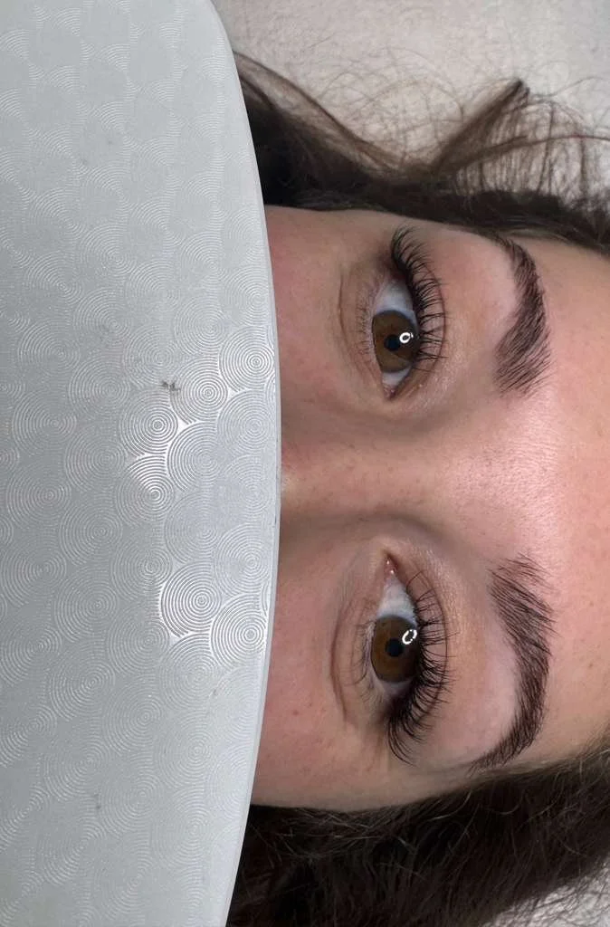 Close-up of a woman lying in bed, with her face partially covered by a white textured blanket, showing her brown eyes, eyelashes, and eyebrows.