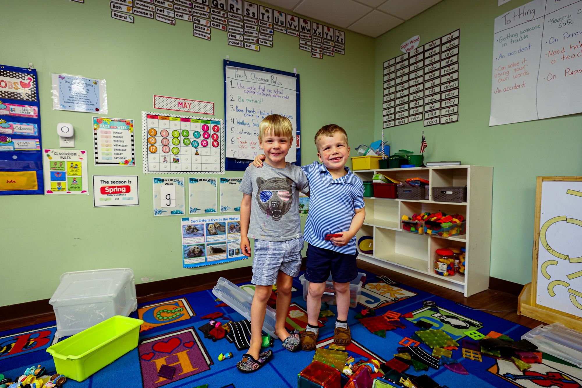 Two young boys standing in a colorful classroom, smiling. The classroom has educational posters and charts on the walls, shelves with toys and supplies, and a bright, engaging environment suitable for young children.