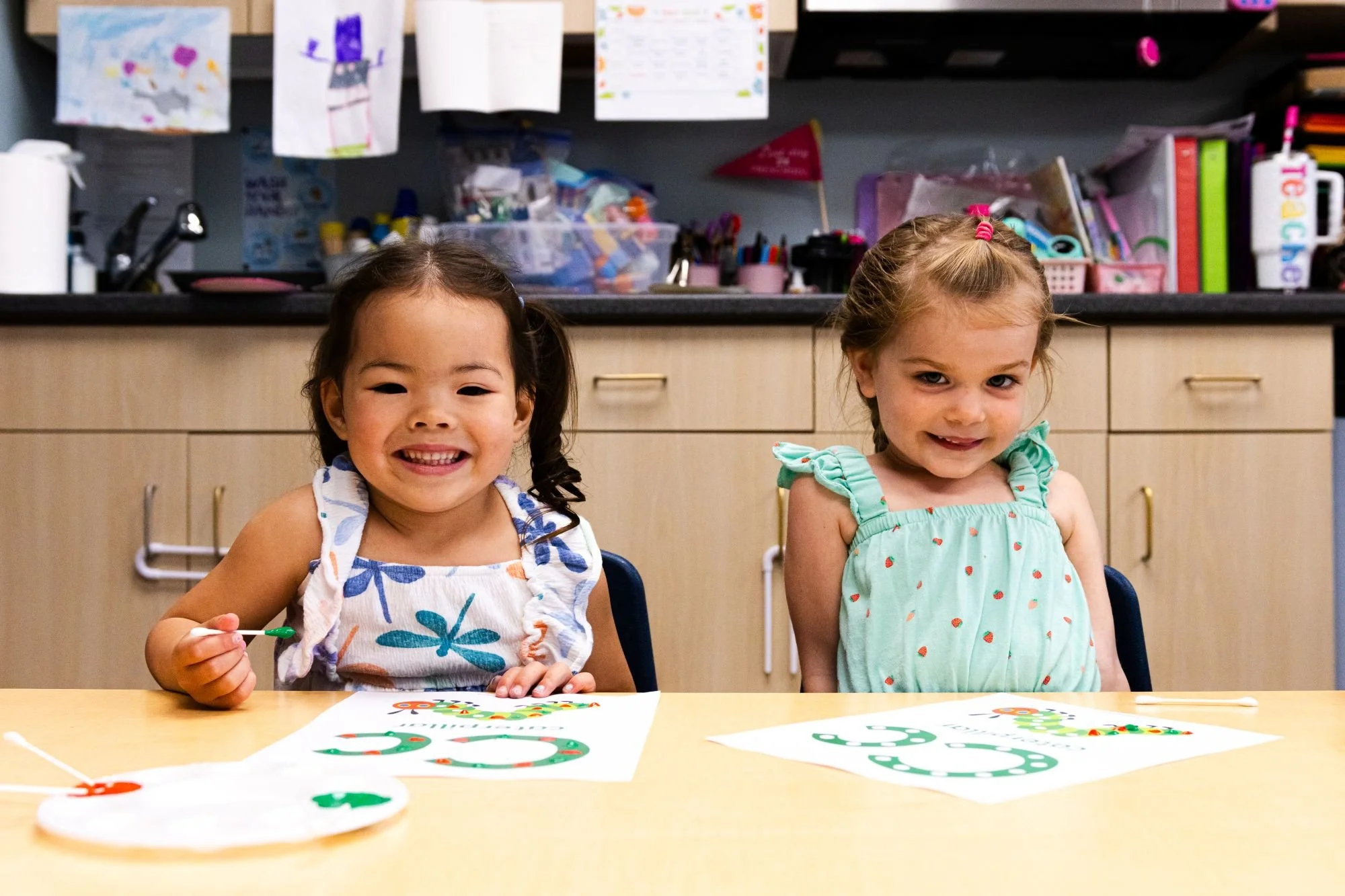 Two young girls sitting at a table with holiday-themed craft projects, smiling at the camera. The girl on the left has dark hair in pigtails, the girl on the right has blonde hair in a ponytail. The table has Christmas wreath cutouts and drawings.