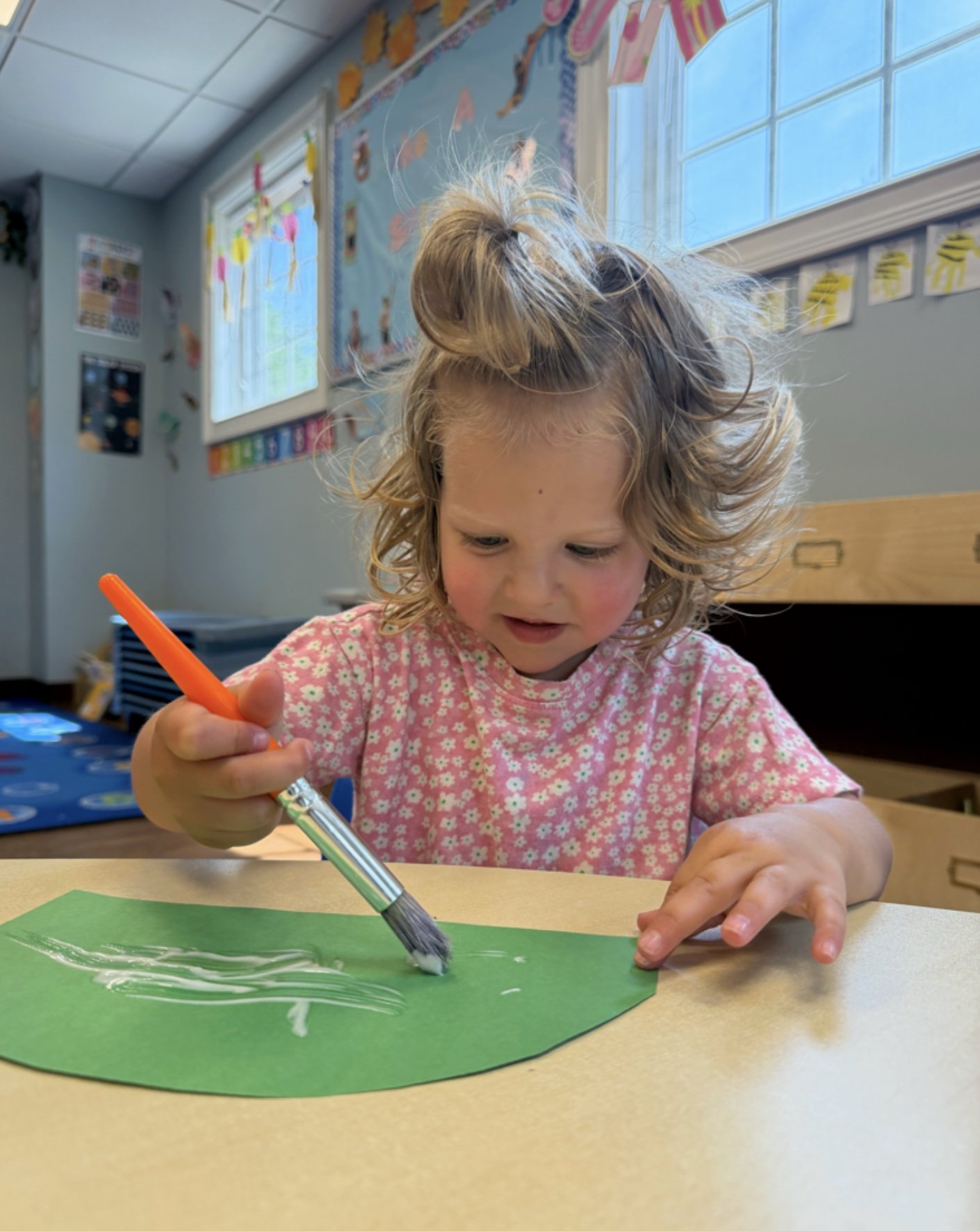 A young girl with curly blonde hair and a pink floral shirt drawing on green paper with white paint using a large brush in a classroom decorated with colorful artwork and educational posters.