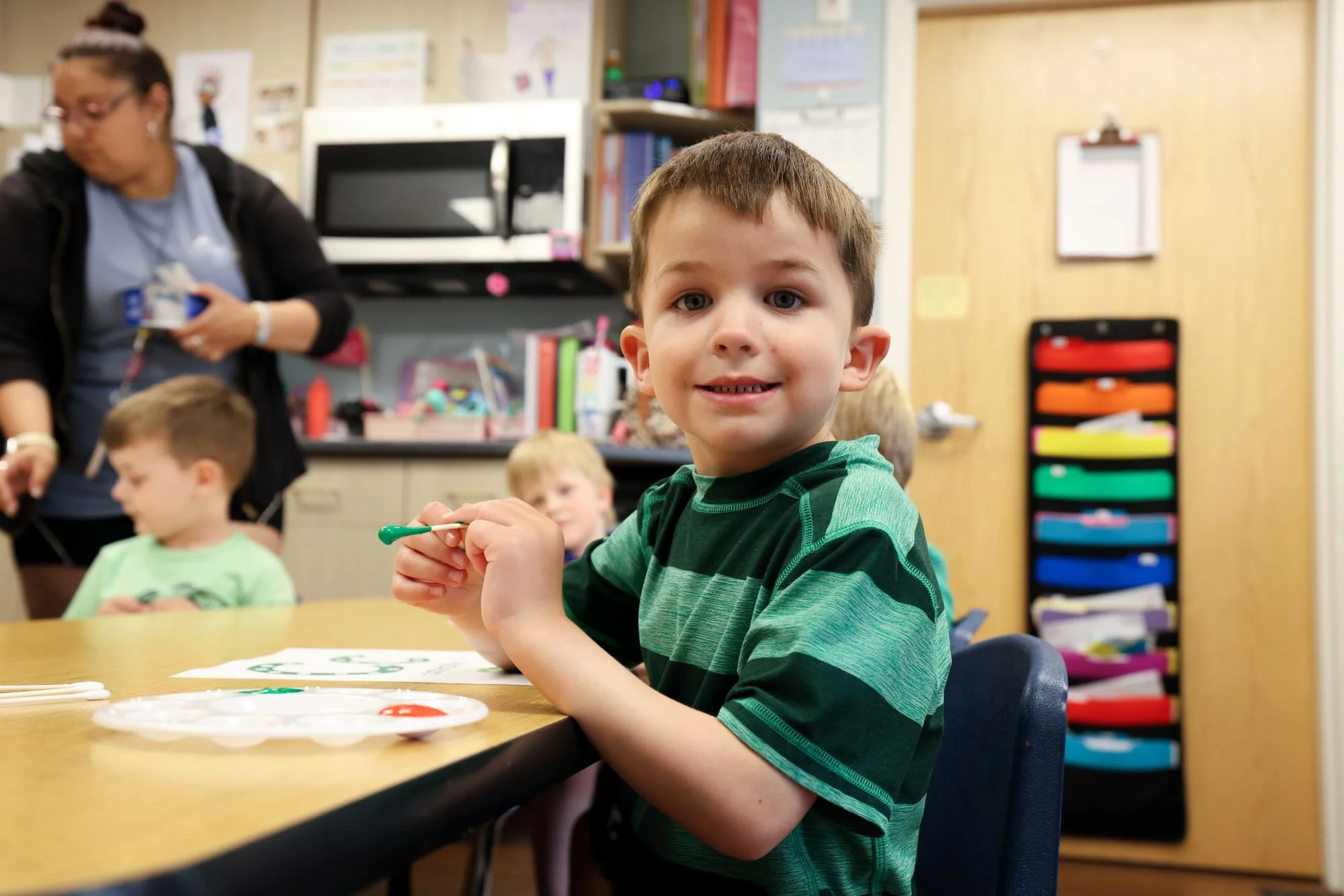 A young boy with brown hair and blue eyes smiles at the camera while sitting at a table with art supplies in a classroom. Other children and a teacher are in the background.