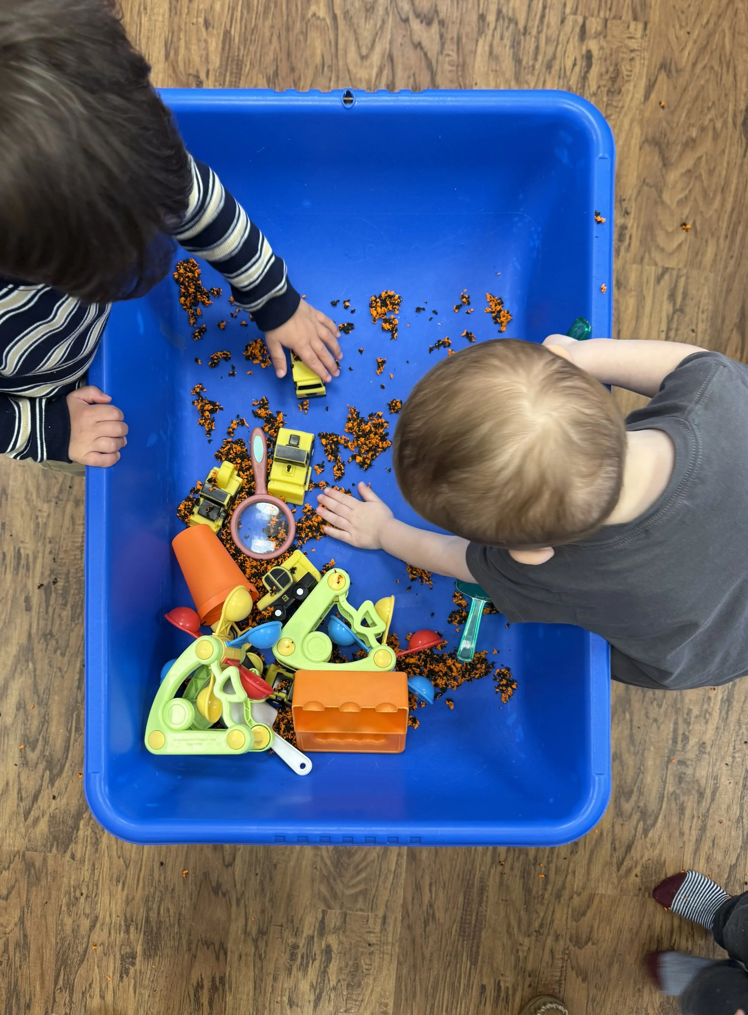 Two children playing with toy construction vehicles and small orange and black objects on a blue plastic table. The children are partially visible, one wearing a striped shirt and the other a solid gray shirt, standing near the table on a wooden floo