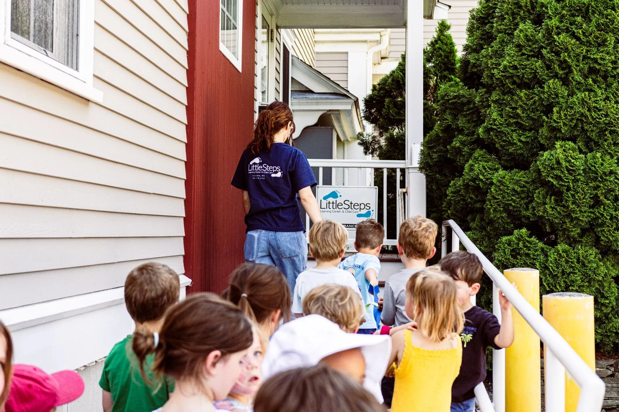 A group of children sitting outside on a railing, listening to a woman in a navy T-shirt with "Little Steps" written on the back, near a building with beige and red siding, surrounded by green trees.