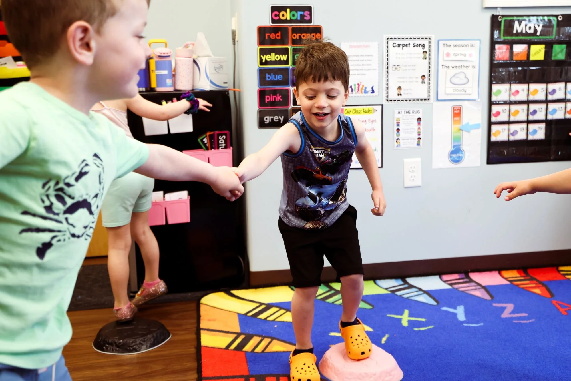 Two young boys holding hands and playing together in a classroom. One boy is wearing a Star Wars shirt and orange Crocs, standing on a pink ball on a colorful rug, while the other boy, in a light green shirt, is partially visible.