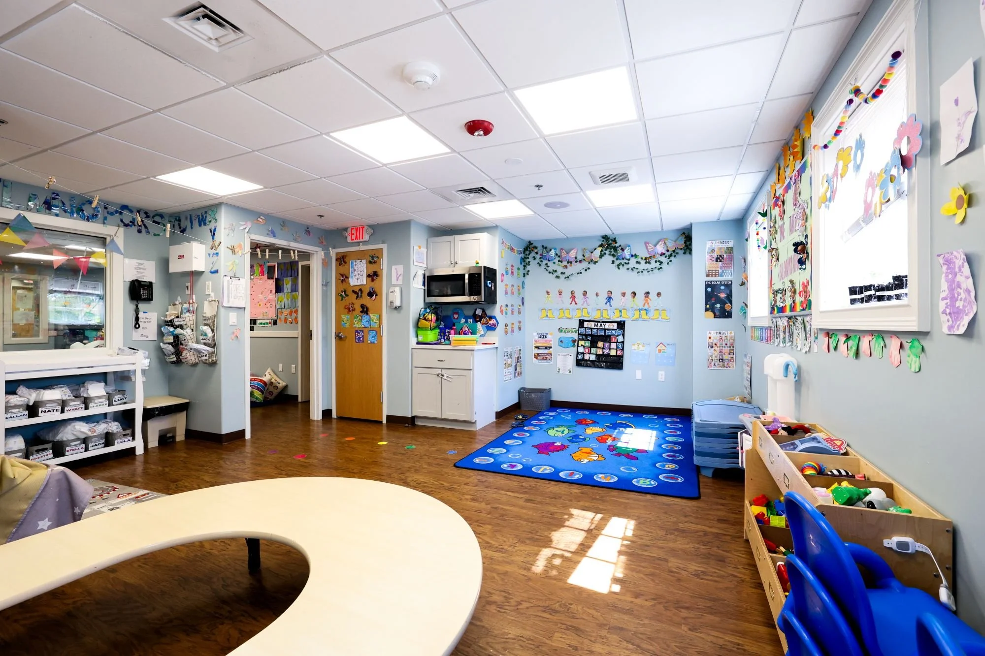 A colorful kindergarten classroom decorated with butterfly and flower cutouts, a blue rug with fish and numbers, shelves with toys, educational posters, and window decorations.