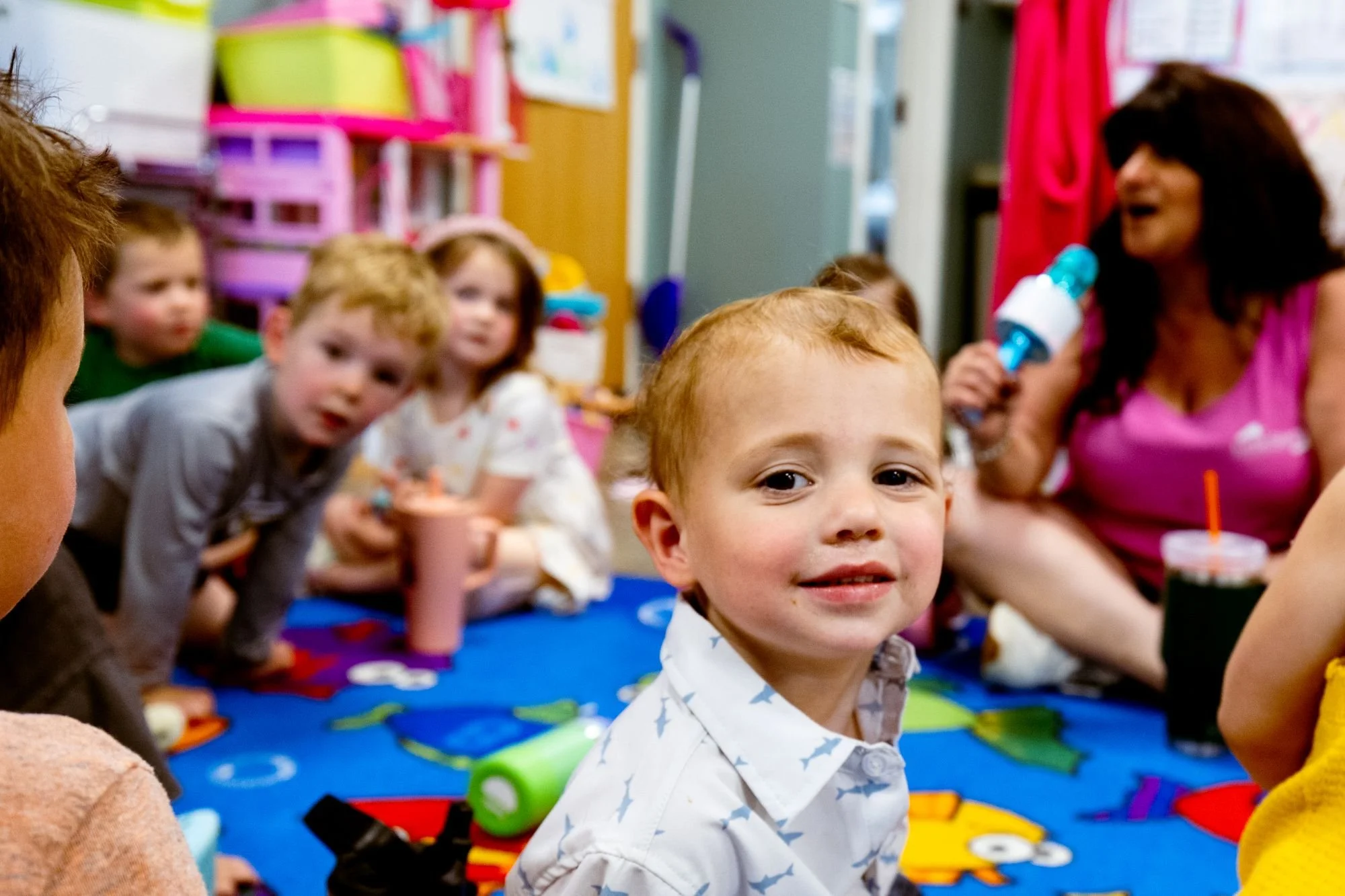 Group of young children and a woman in a colorful classroom, engaging in a learning activity on the floor with toys and colorful decorations.