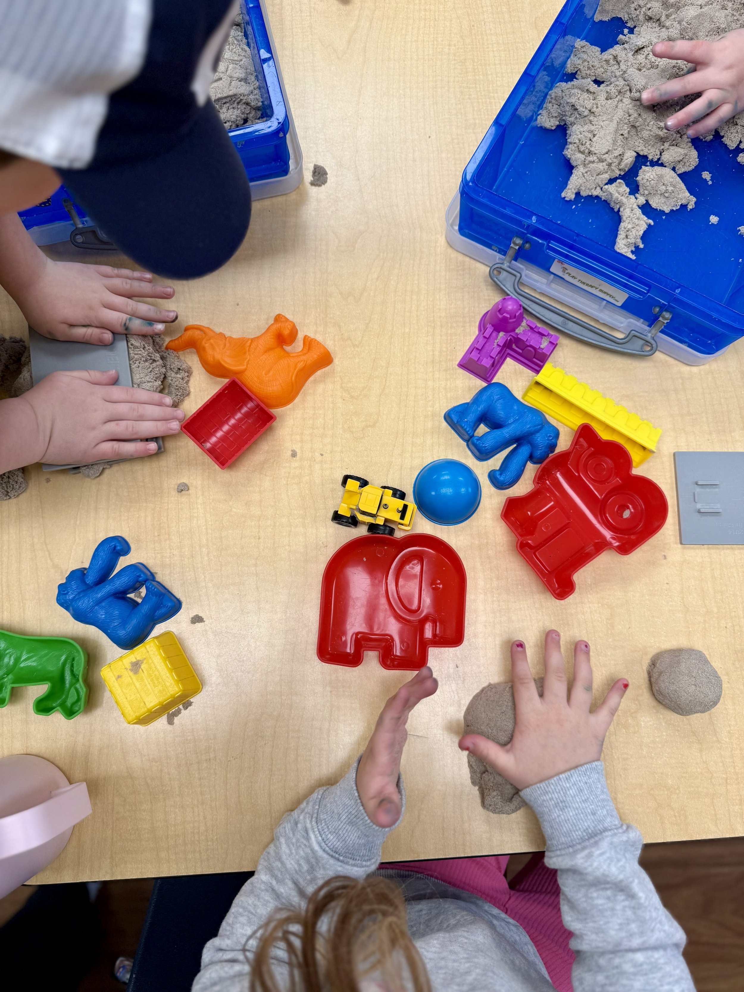Children playing with kinetic sand and colorful plastic toys on a wooden table.