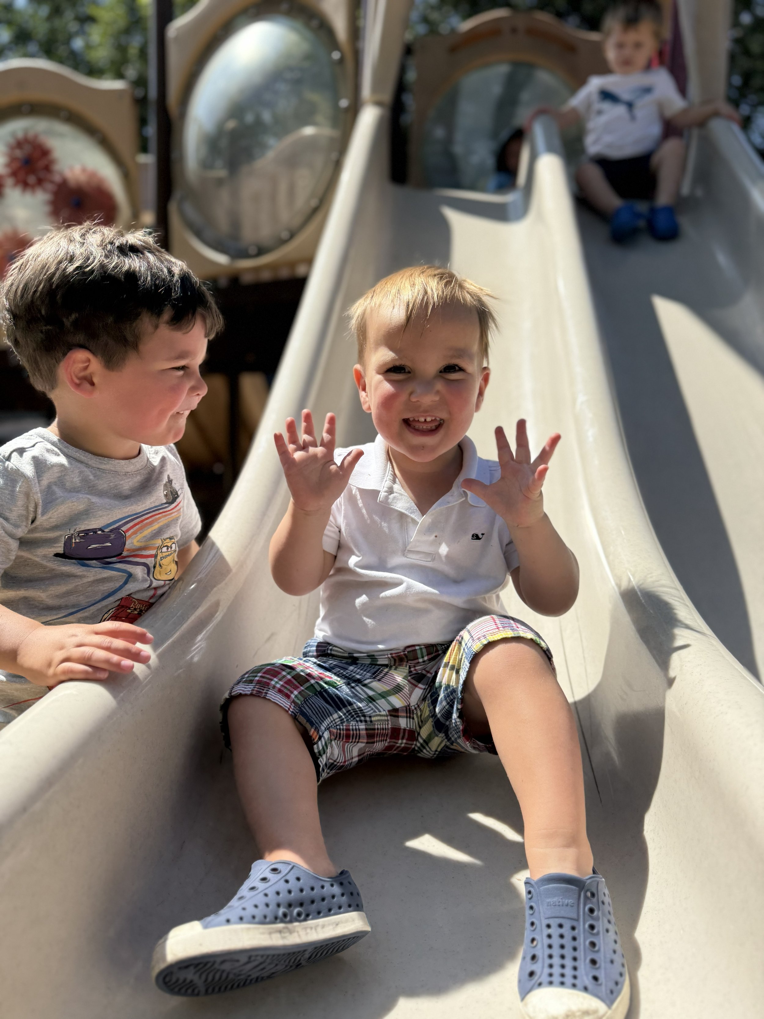 Three young children playing on a slide outdoors in a park or playground area. One child is sitting at the bottom, smiling and waving with their hands raised. Two other children are further up the slide, with one about to reach the bottom and another at the top.