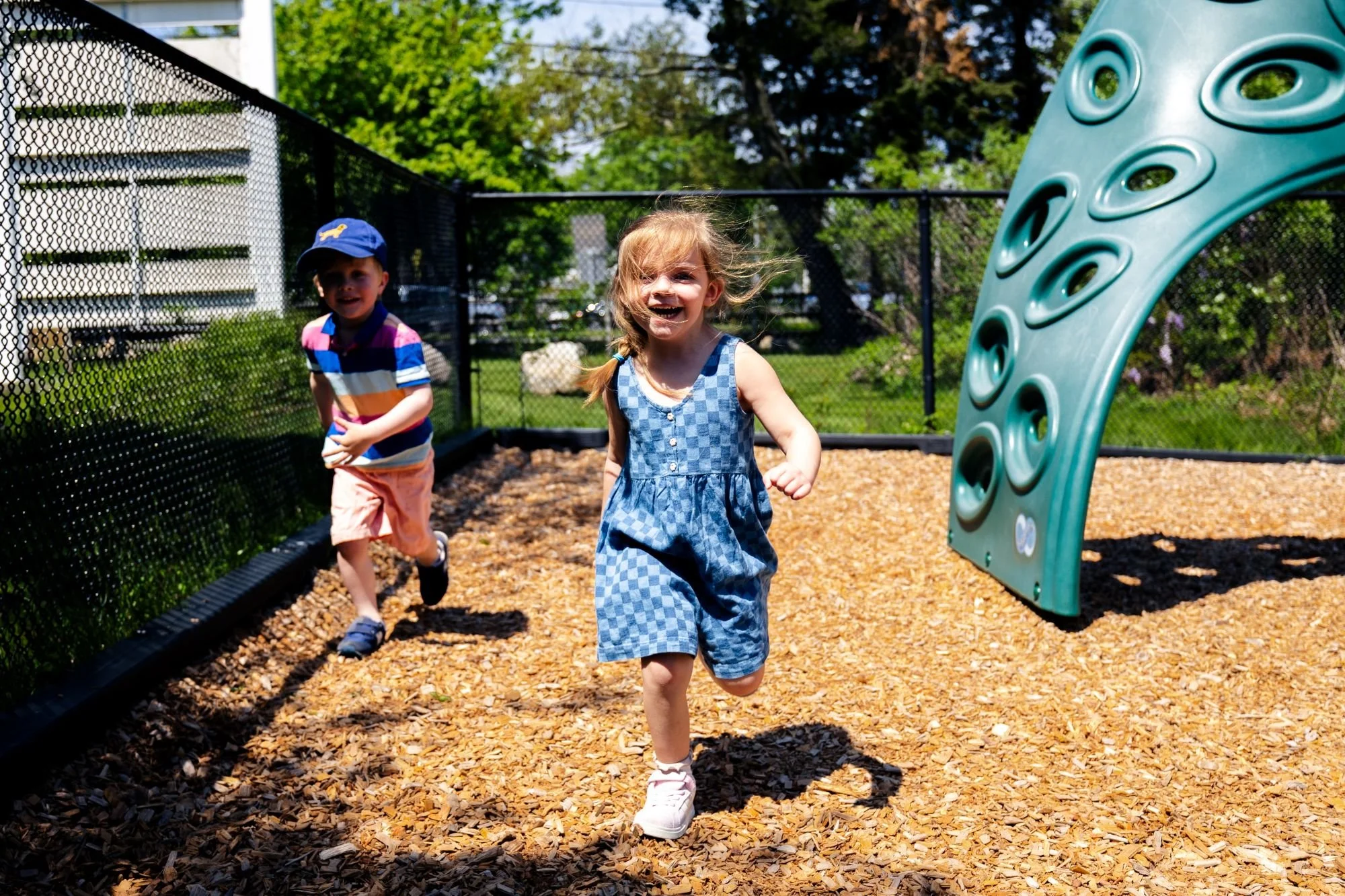 Two kids, a boy wearing a colorful striped shirt and a girl in a blue checkered dress, playing and running at a playground surrounded by a black safety fence on a sunny day with trees in the background.