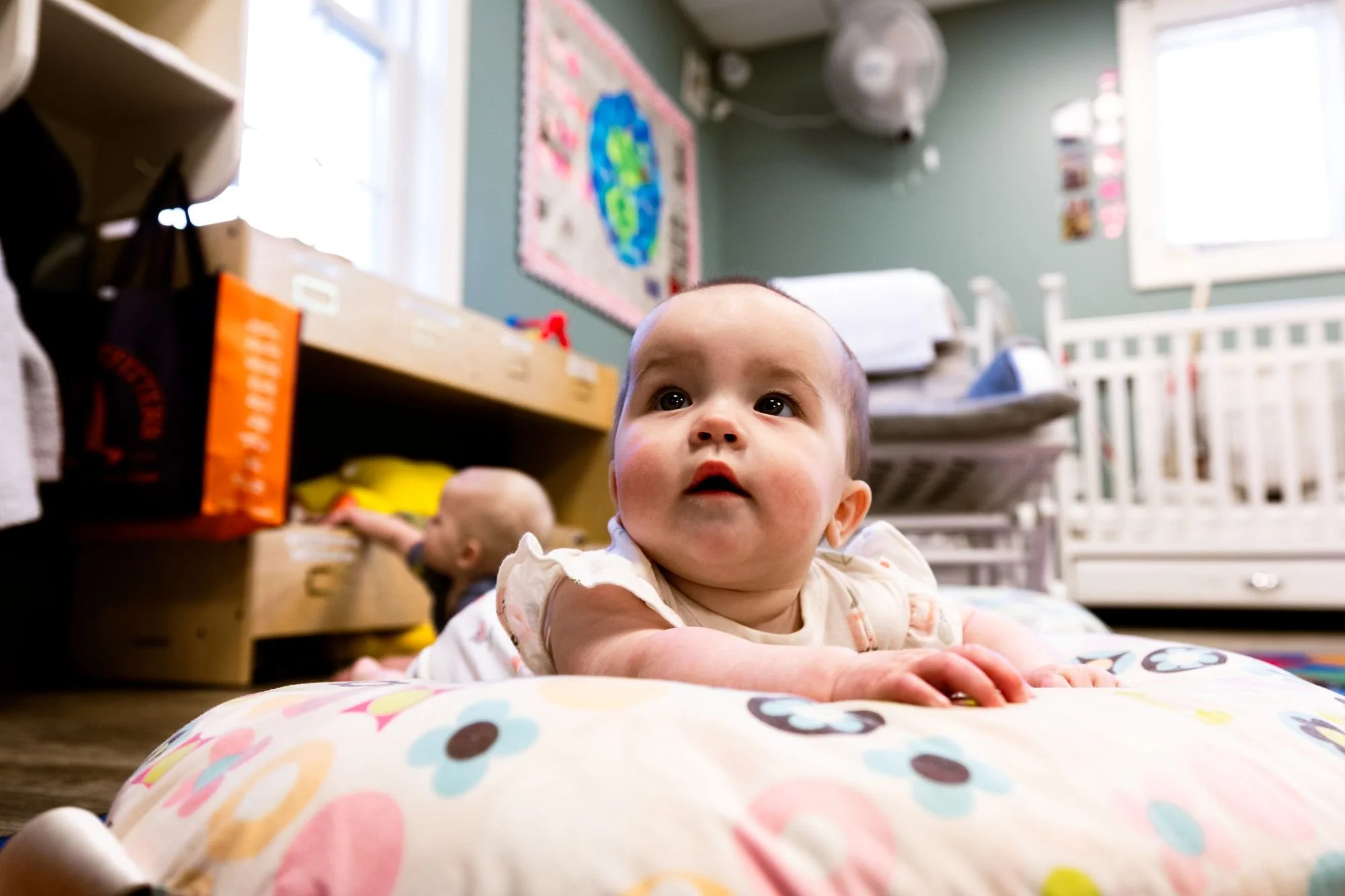 A baby girl with big brown eyes and dark hair lying on her stomach on a colorful polka dot blanket, with a colorful play mat and a toy in the background in a room with green walls and white furniture.