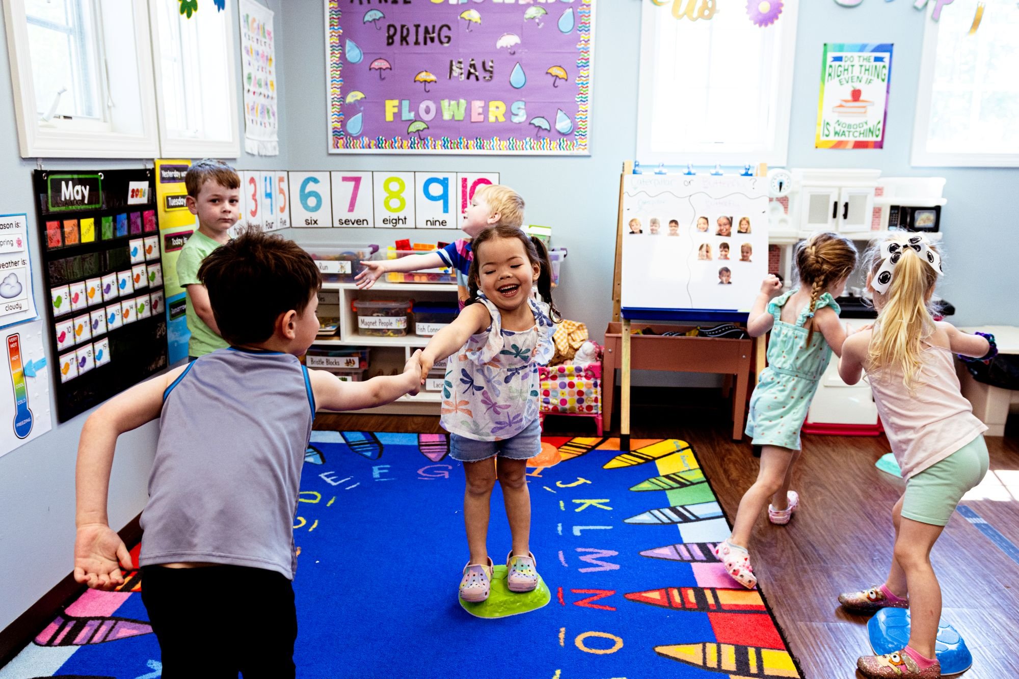 Children playing in a colorful classroom with educational posters on the walls, including numbers, months, and weather, while kids dance and hold hands on a vibrant alphabet rug.