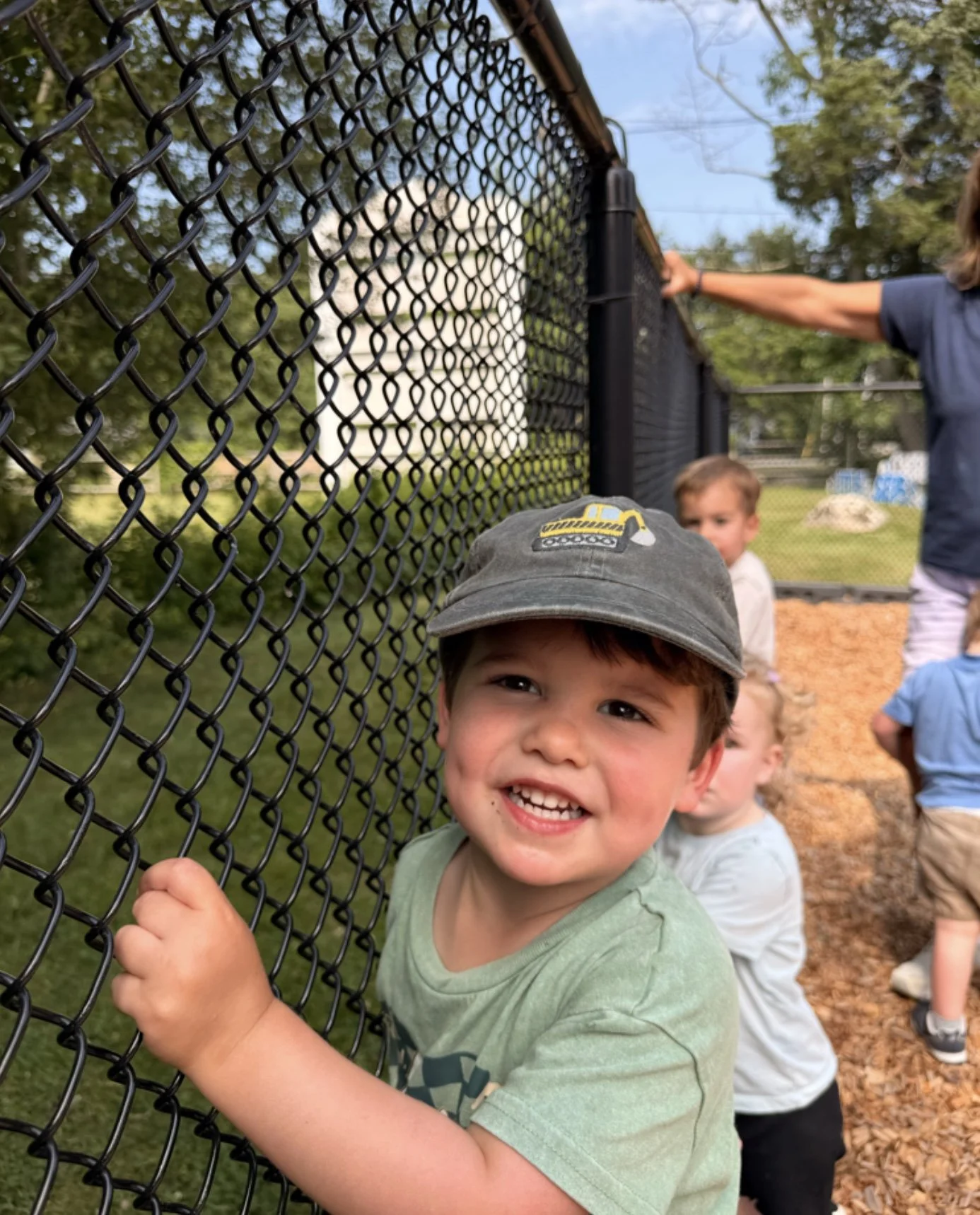 Smiling young boy with a cap holding onto a chain-link fence, with other children in the background.