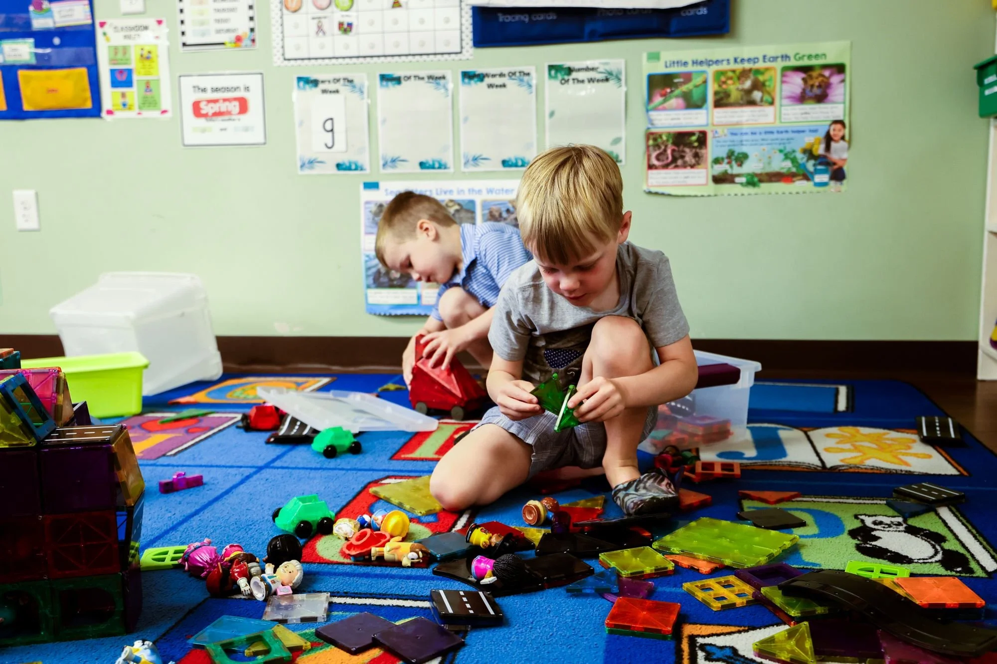 Two young boys sitting on a colorful carpet in a classroom, playing with small toy cars and figurines surrounded by various toys and storage bins.