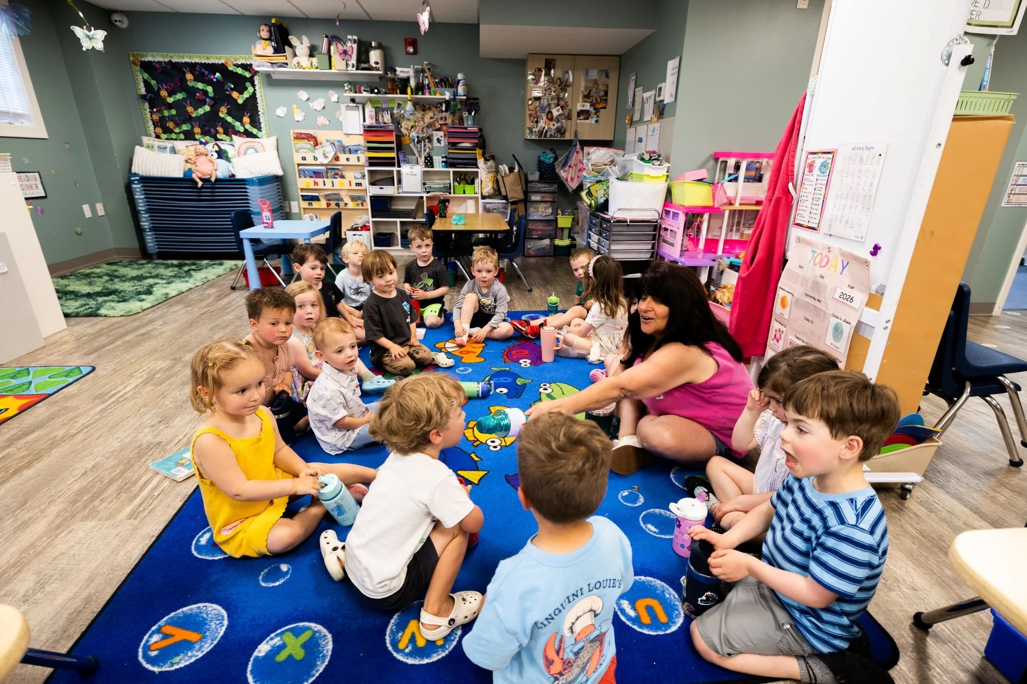 A group of young children sitting on a colorful school carpet in a classroom, engaged in a story or lesson, with a teacher or caregiver leading the activity.