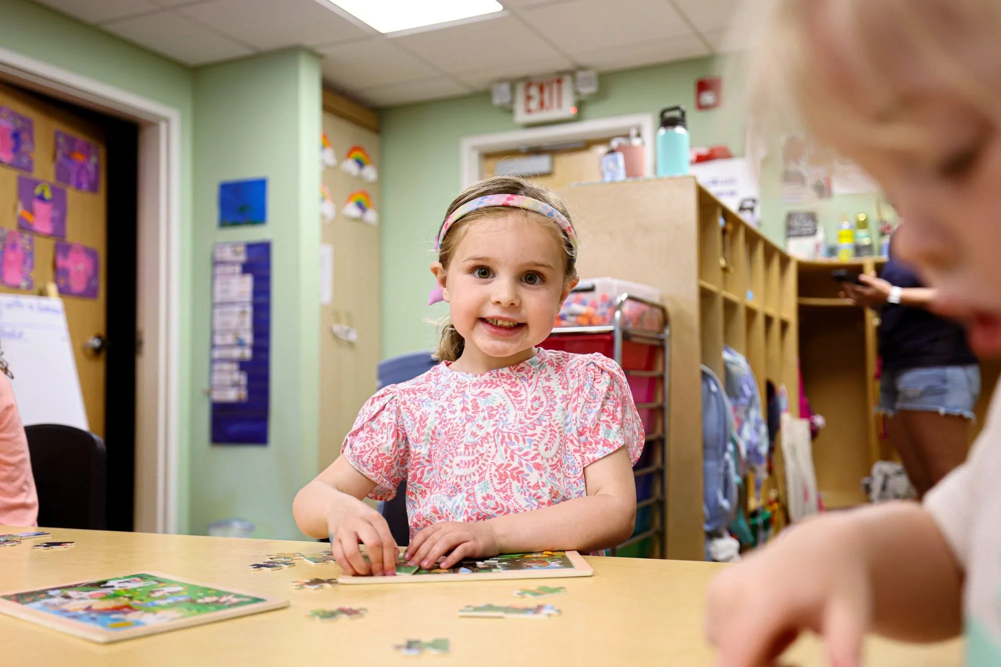 A young girl with a floral headband and bright smile sitting at a table, working on a jigsaw puzzle in a colorful classroom with art on the walls and shelves filled with books and toys.
