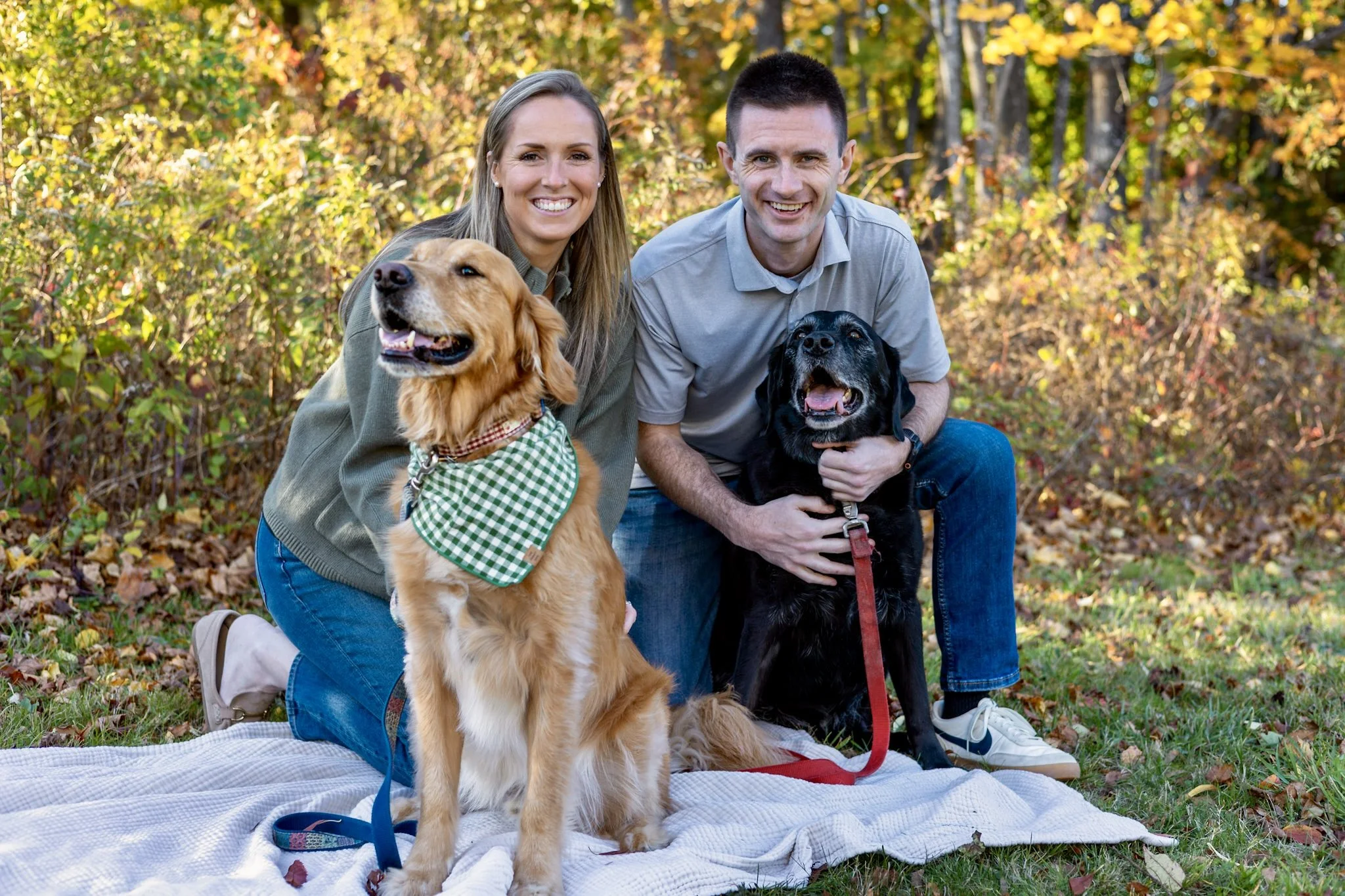 A smiling couple with two dogs, a golden retriever and a black lab, kneeling on a blanket in a park with autumn foliage.