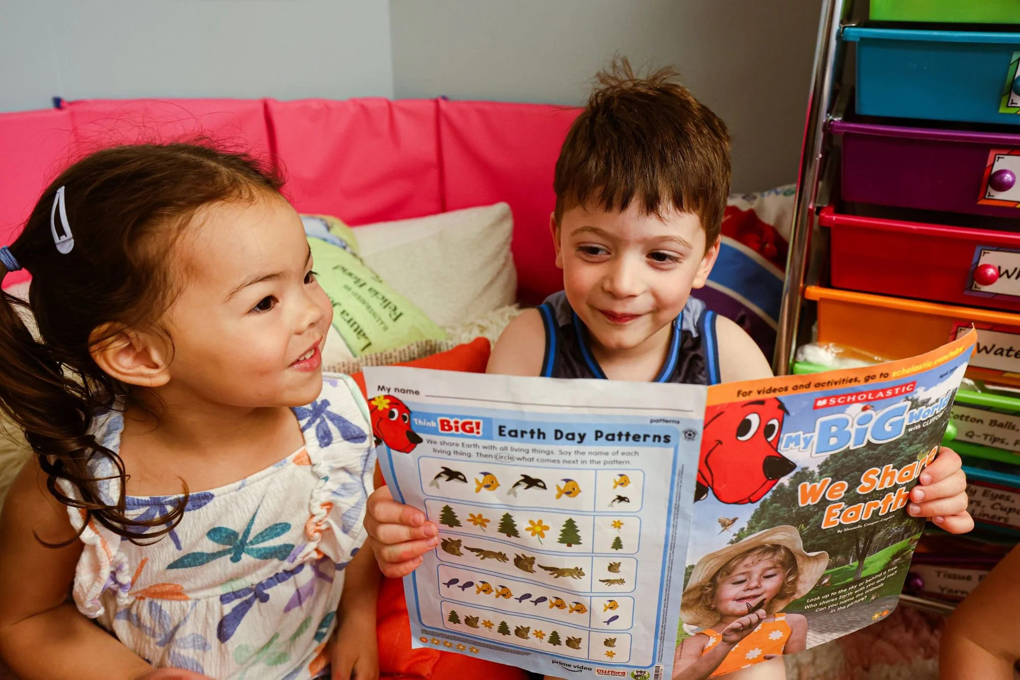 Two young children, a girl and a boy, are sitting together reading a colorful book titled 'My Big Earth Day'. The girl has dark hair tied in pigtails with white hair clips and wears a white dress with colorful flower patterns. The boy has light brown