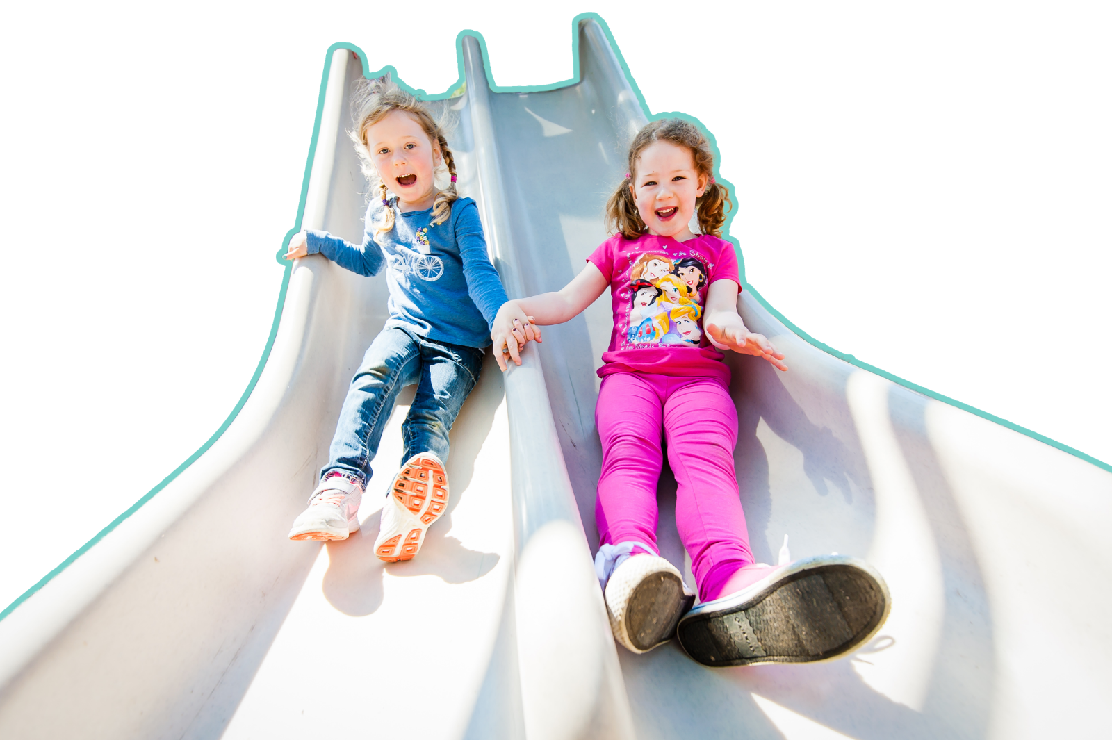 Two young girls sliding down a playground slide, holding hands and smiling. One girl is wearing a pink shirt with cartoon characters and pink pants, the other girl is wearing a blue shirt with jeans.