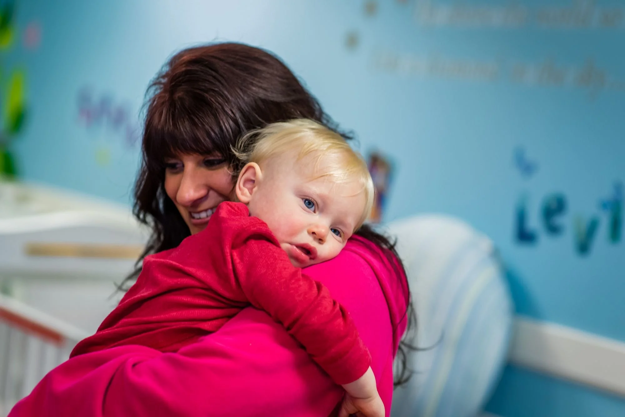 A woman with dark hair holding a young blonde-haired child in a hospital or clinic setting.