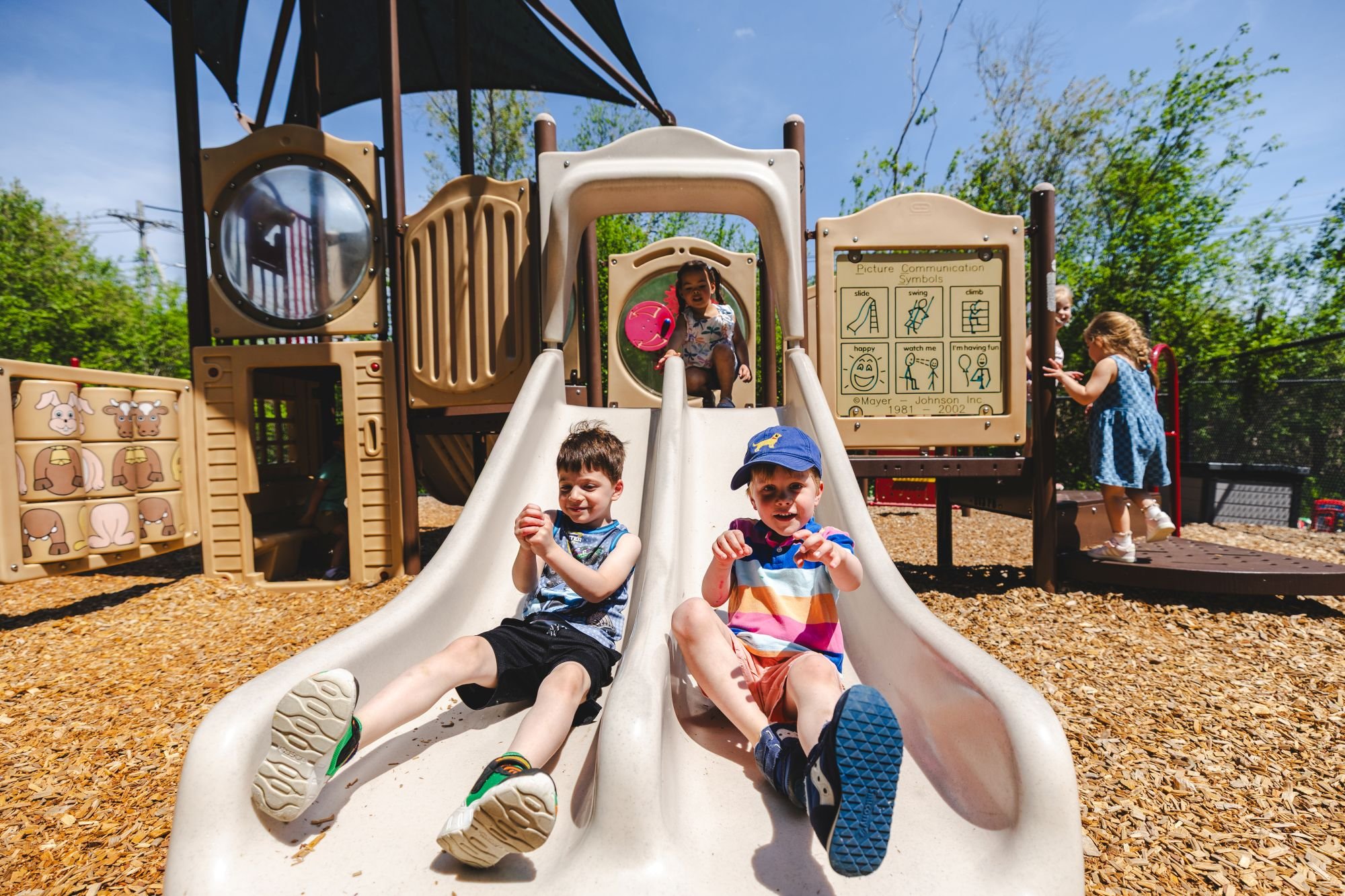 Children playing on a playground slide outdoors, with trees and a clear blue sky in the background. Some children are sliding down, and others are standing or sitting around the playground.