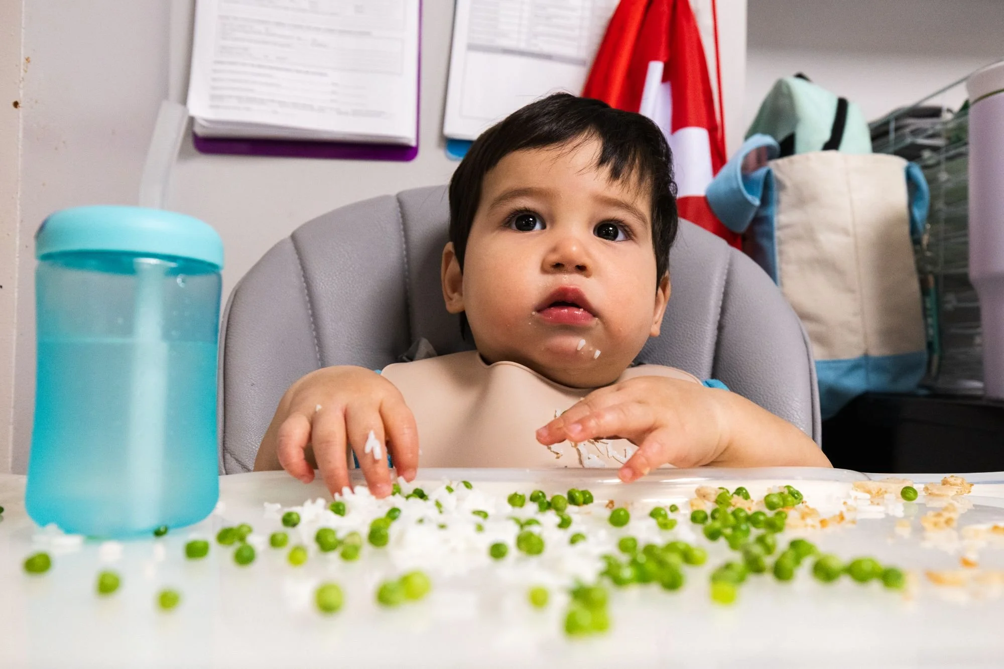 A young child with dark hair and big eyes sitting in a high chair, eating a meal with peas and other foods on a white tray. There is a blue water bottle to the left, and the background shows various papers, a red and white cloth, and a laundry basket