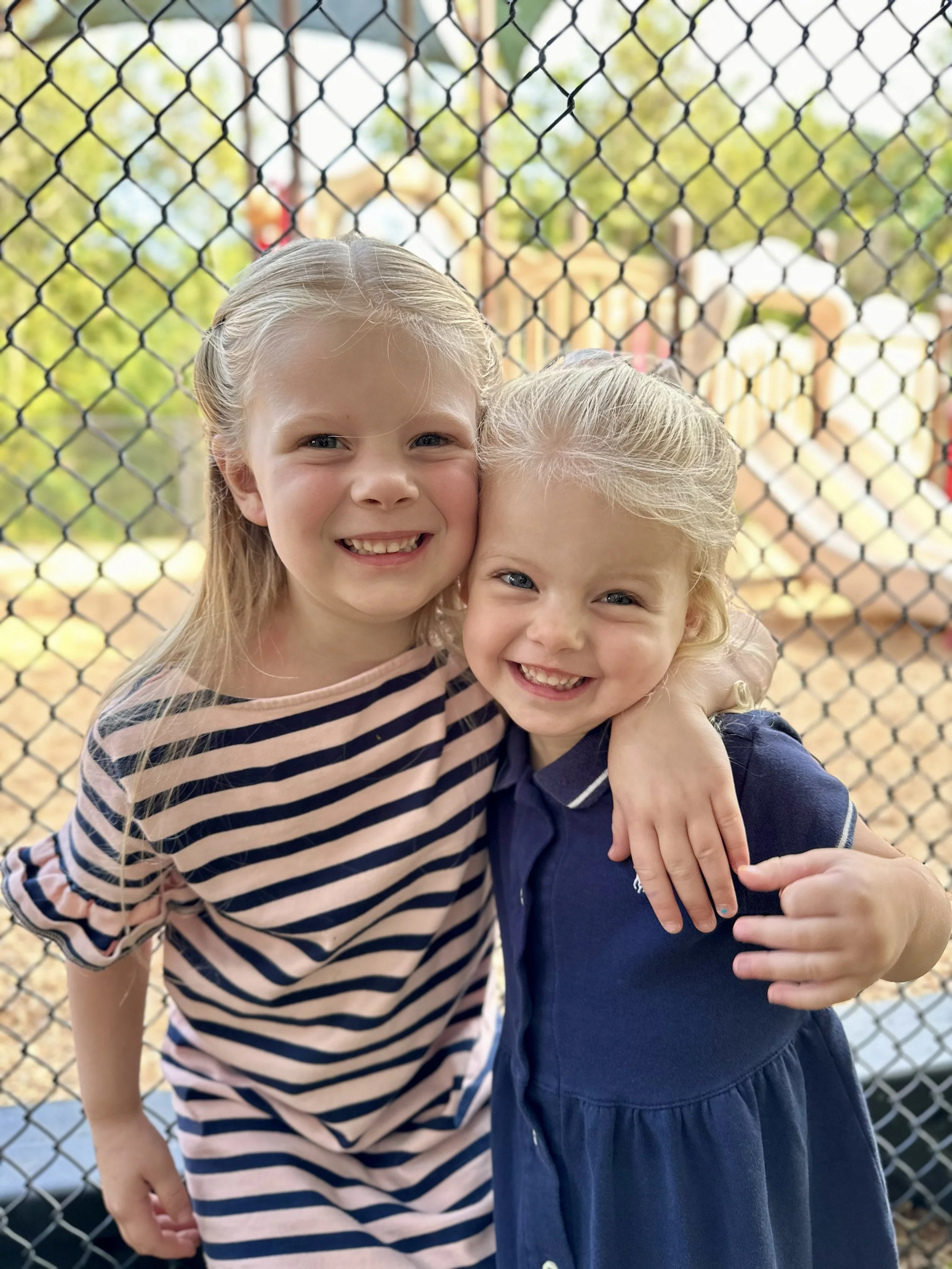 Two young girls smiling and hugging each other in front of a chain-link fence at a playground, outdoors on a sunny day.