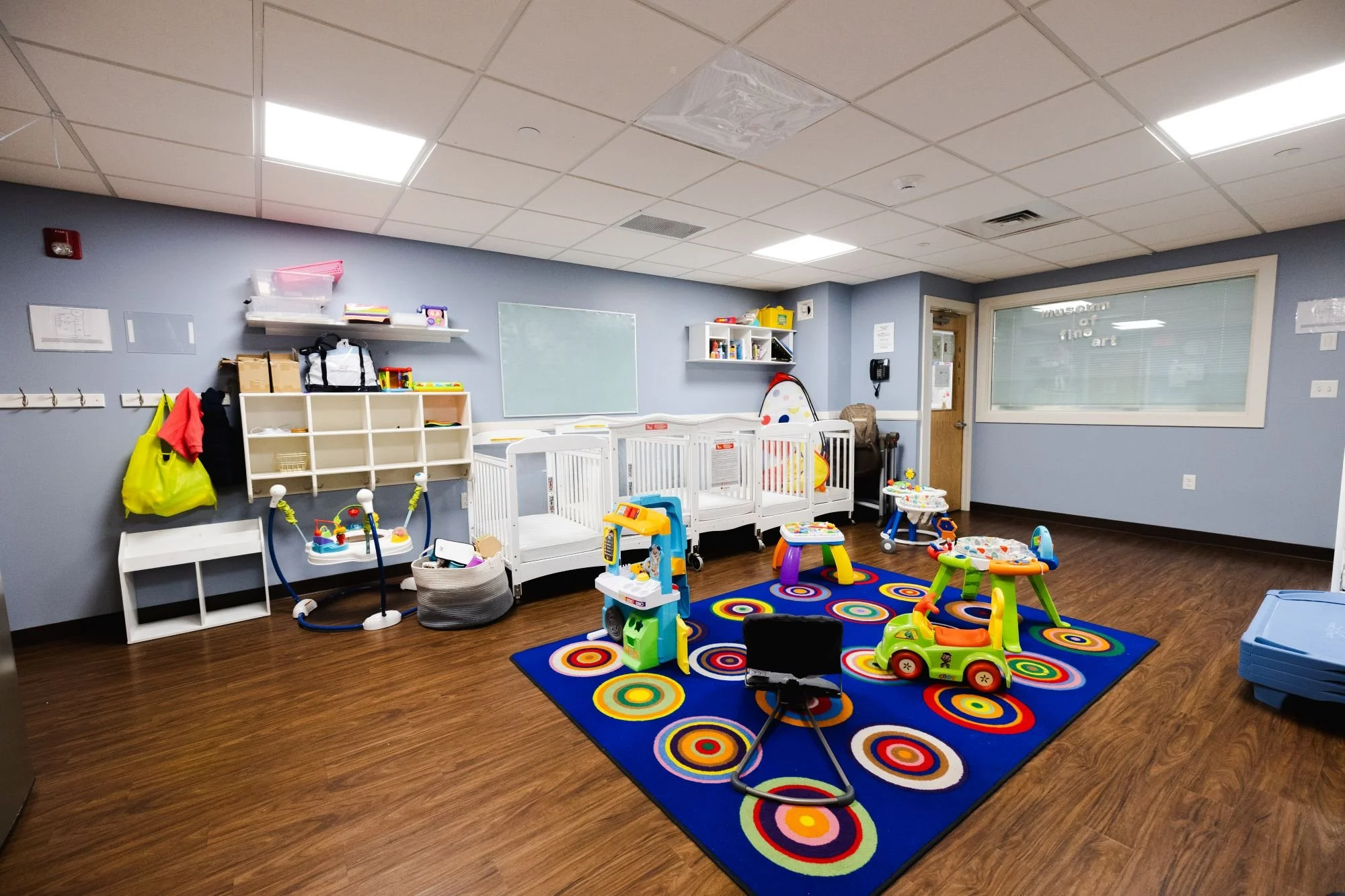 A pediatric playroom with colorful toys, a rainbow rug, a white playpen, and organizational shelves against a light blue wall.