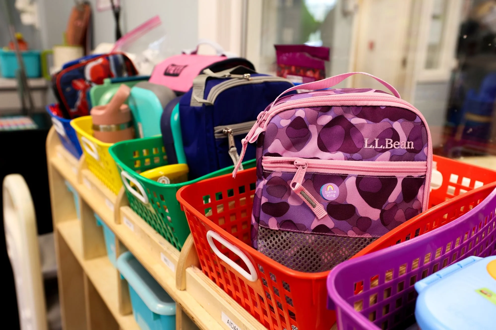 Colorful classroom supplies including multiple school backpacks, water bottles, and storage containers placed on a wooden shelf in a school supplies aisle.