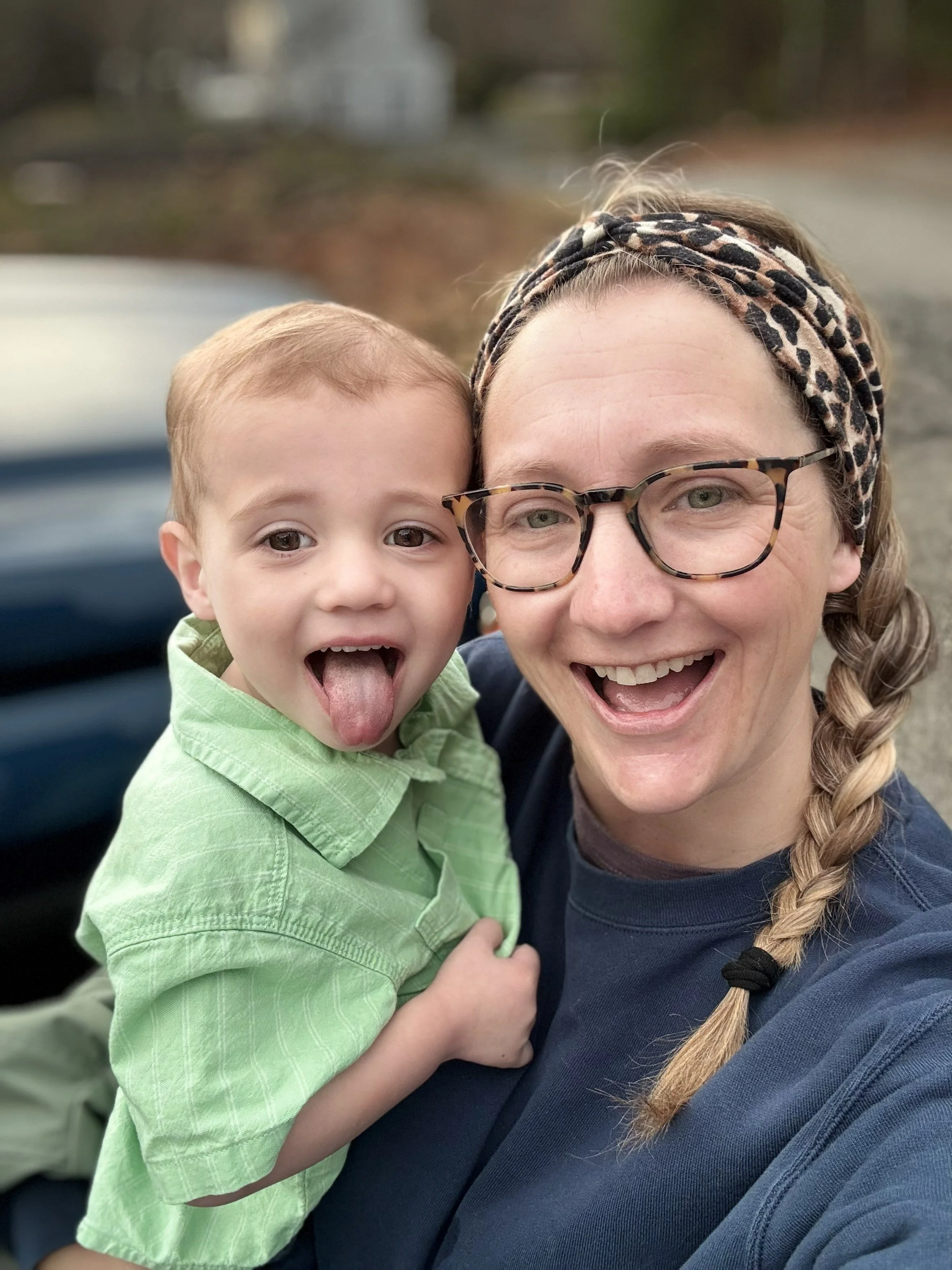 A woman and young boy smiling and laughing outdoors on a cloudy day, close-up selfie with blurred background.