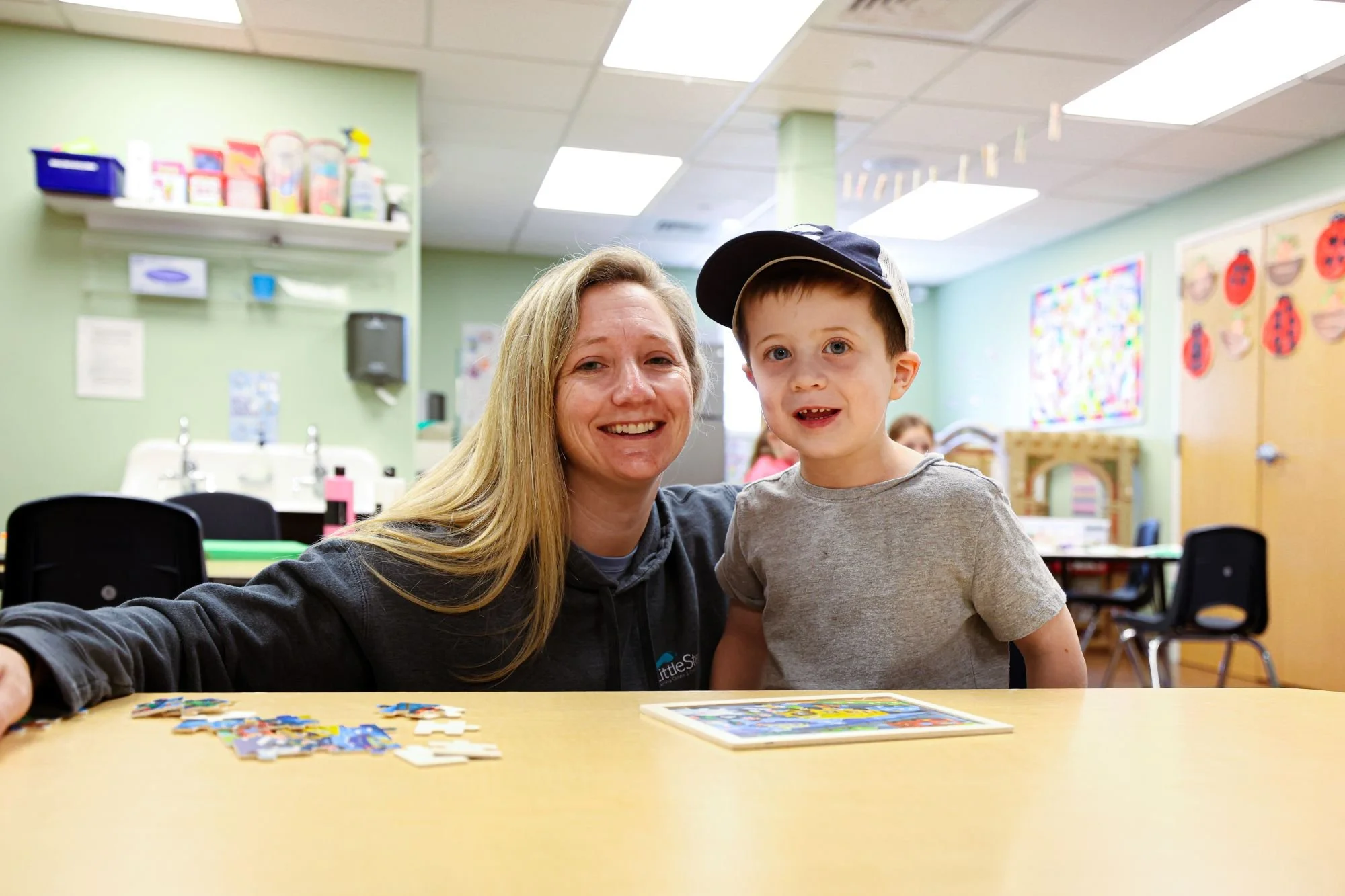 A woman and a young boy are sitting at a table in a classroom, smiling at the camera. The table has puzzles and a board game on it. The classroom walls are decorated with colorful artwork, and there are chairs and shelves in the background.