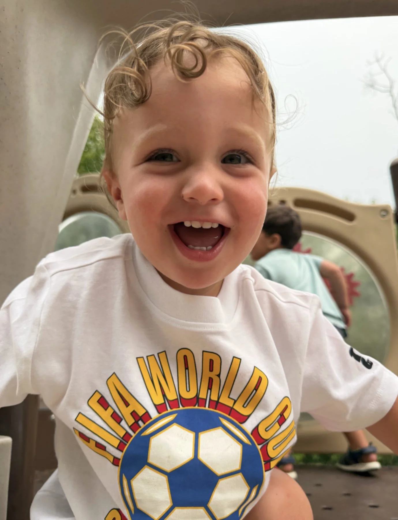 A young boy with curly blonde hair smiling indoors, wearing a white T-shirt with a colorful soccer ball graphic and the words "FA World Cup".