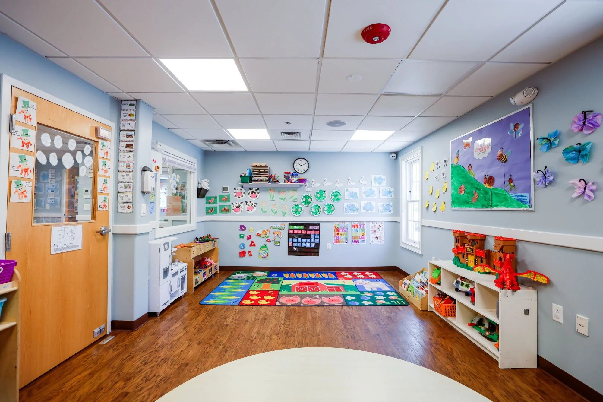 A colorful, tidy preschool classroom with wall decorations, a fire alarm on the ceiling, and a play area with educational toys and a rug with animal and number patterns.