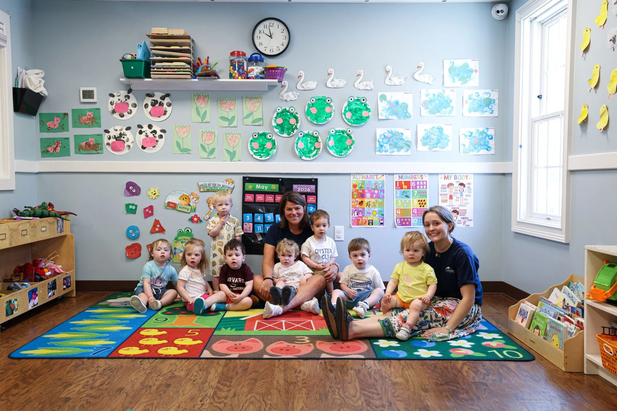 A group of young children and two adult women sitting on a colorful educational rug in a bright classroom. The classroom walls are decorated with children's artwork, including animal and nature-themed paintings, and educational posters about the alphabet, numbers, and body parts. Shelves with toys and books are visible, and sunlight enters through a window on the right.