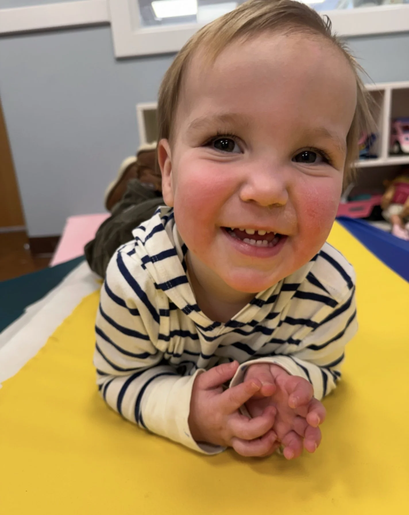 A young boy with light brown hair and a wide smile, lying on his stomach on a yellow surface, wearing a striped long-sleeve shirt. The background shows a room with a light blue wall and shelves with toys.