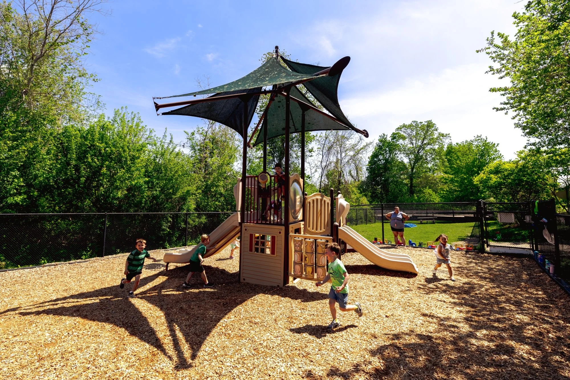 Children playing on a playground with a slide, climbing structure, and shade canopy, surrounded by trees and a fence.