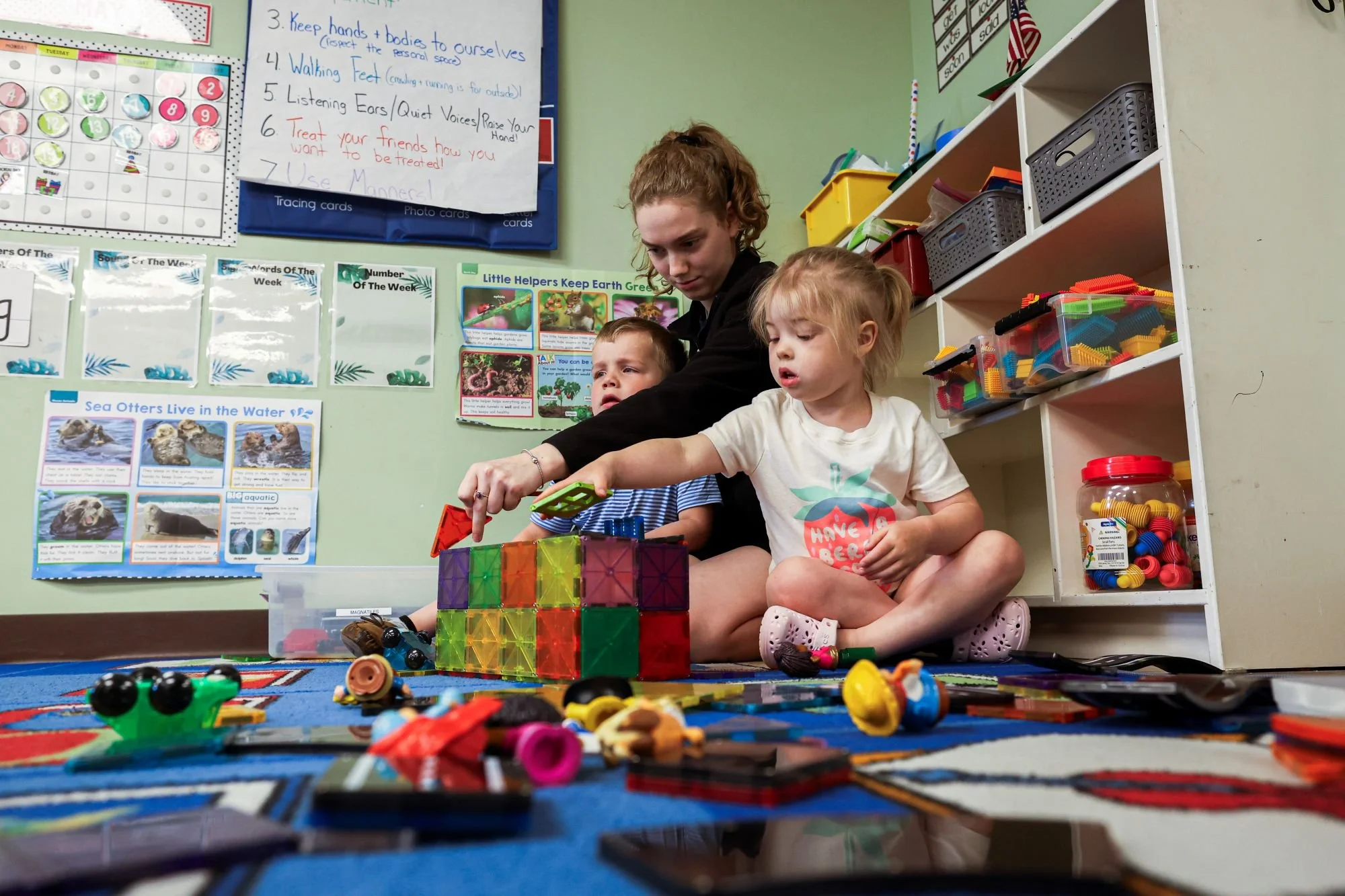 Two young children, one girl and one boy, sitting on the floor and playing with colorful building blocks and toys in a classroom. An older girl is leaning over them, helping or showing something. The classroom walls are decorated with educational pos