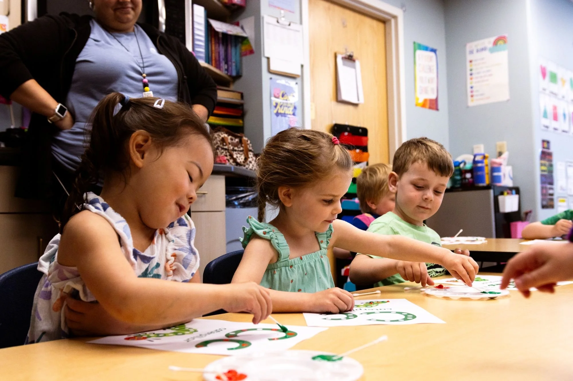 Four young children sitting at a table engaged in an arts and crafts activity, with a teacher or adult observing in the background in a classroom decorated with colorful posters and supplies.