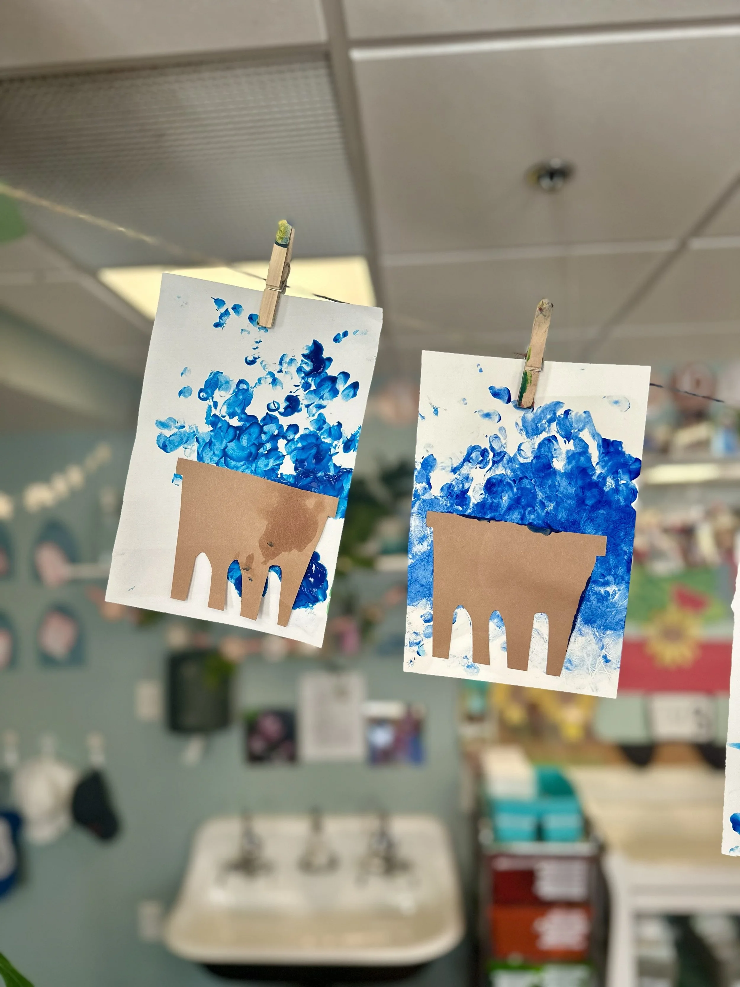 Two photos of paintings of ice cream cones with blue and white brushstrokes, hanging on a line with clothespins in a classroom or art space.