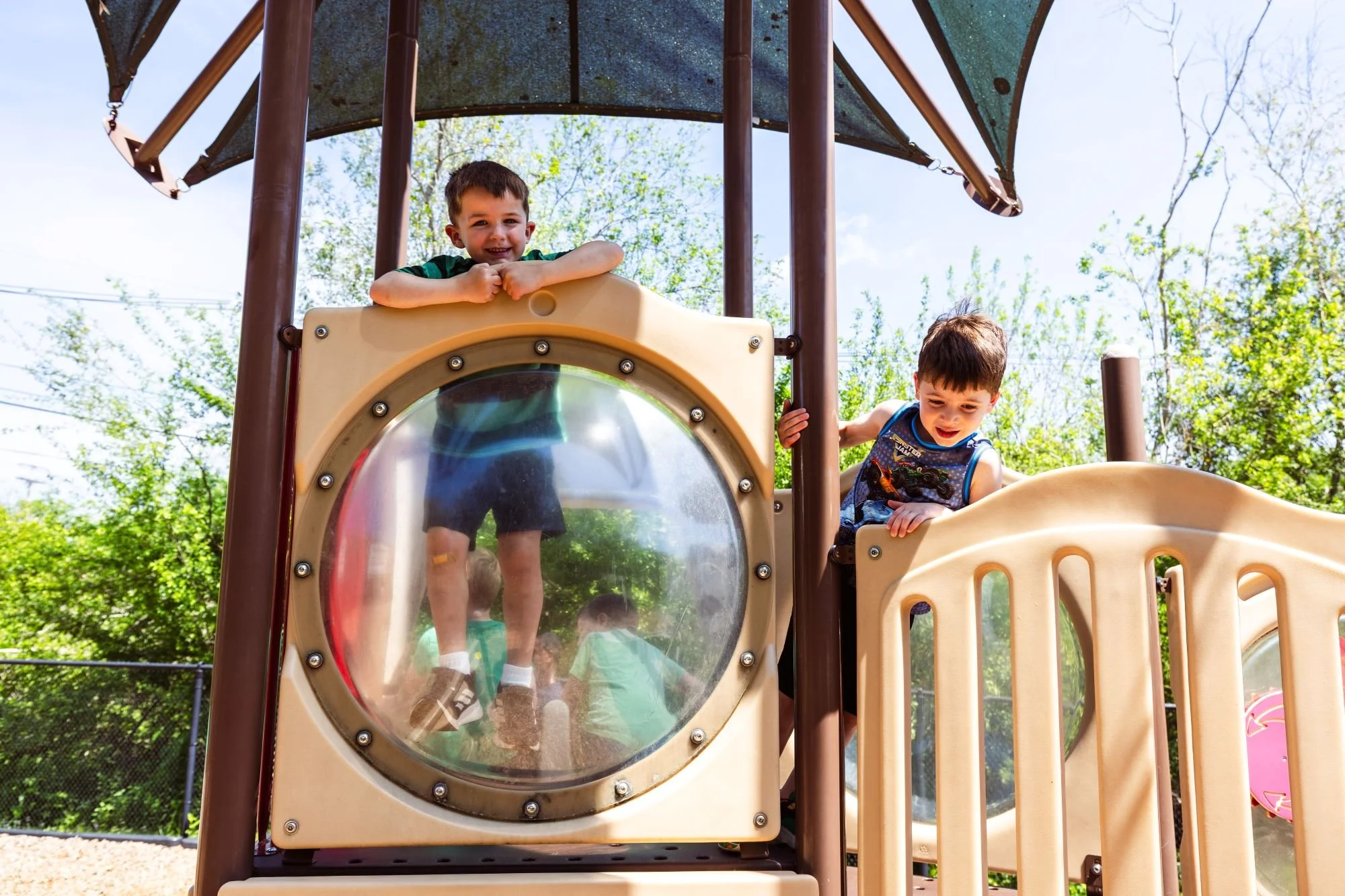Two young boys playing on a playground structure outdoors on a sunny day. One boy is standing behind a transparent circular window, smiling and leaning on a beige panel, while the other is climbing or holding onto the side of the structure, looking d