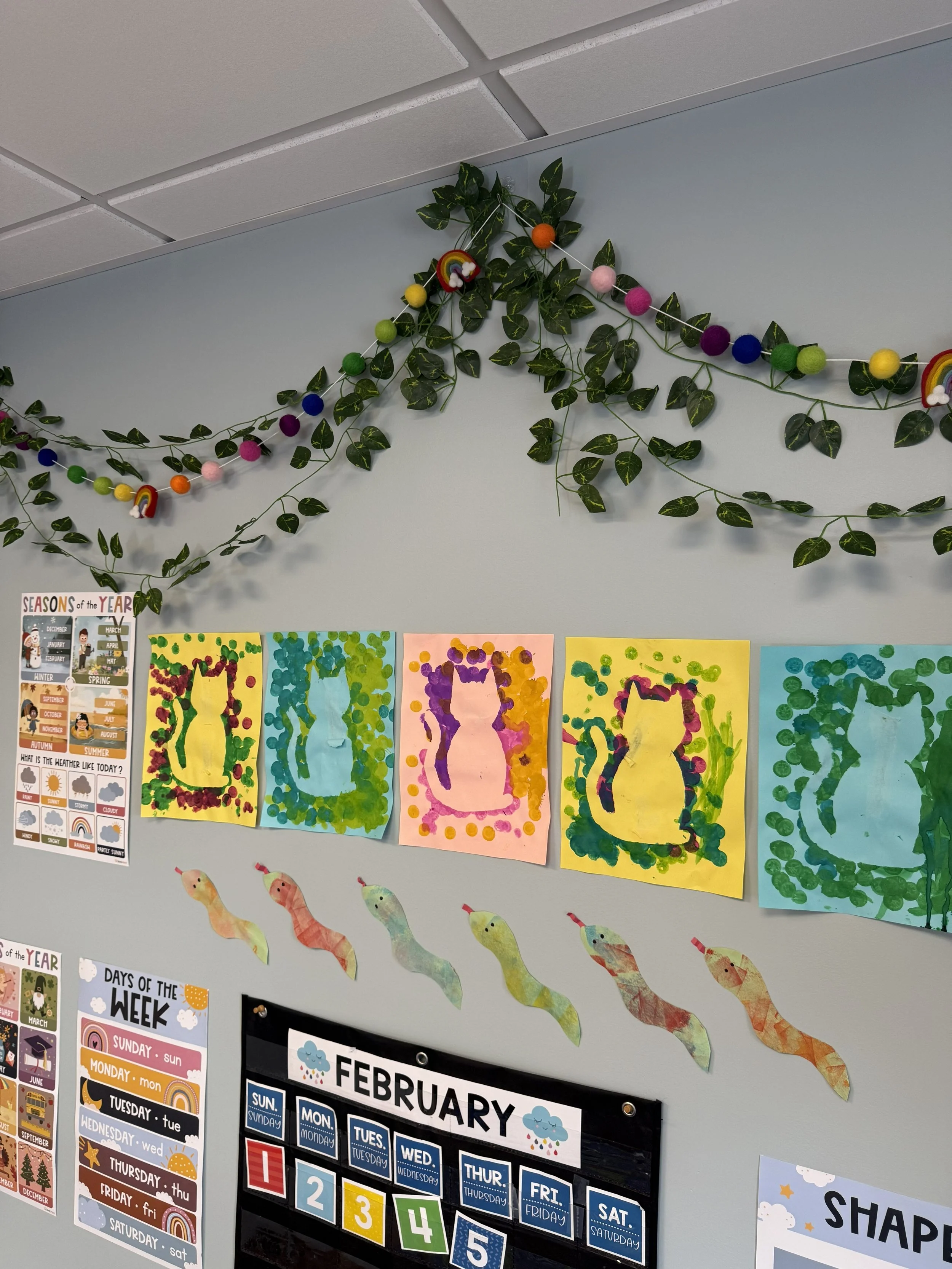Decorative wall with colorful paper art and educational posters featuring seasons, weather, and the month of February, along with paper snake craft at the bottom.