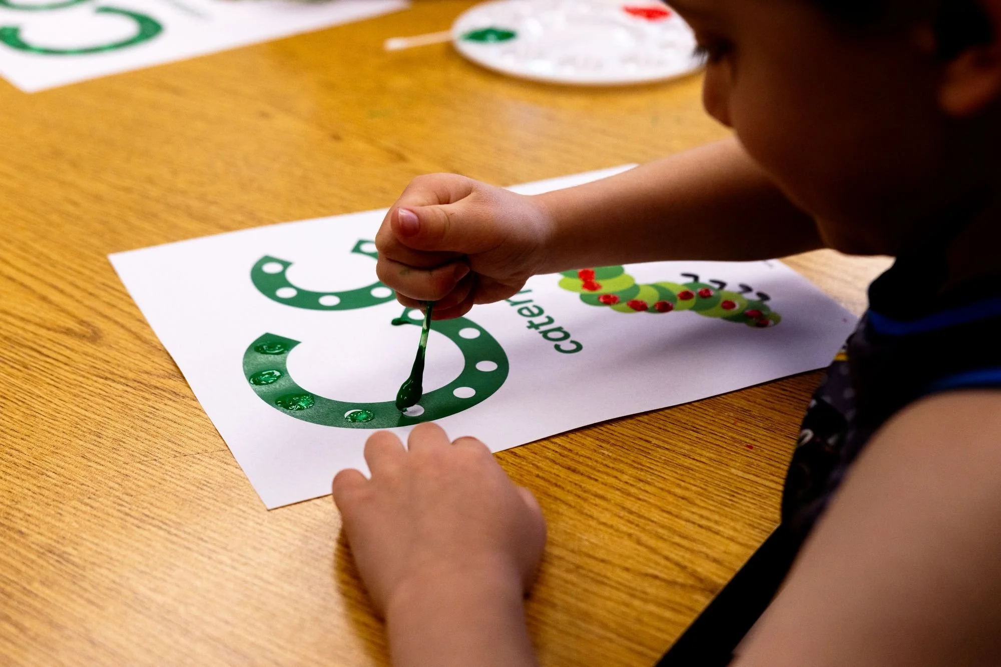A child is using a paintbrush to add green dots to a Christmas-themed Christmas coloring page on a wooden table.