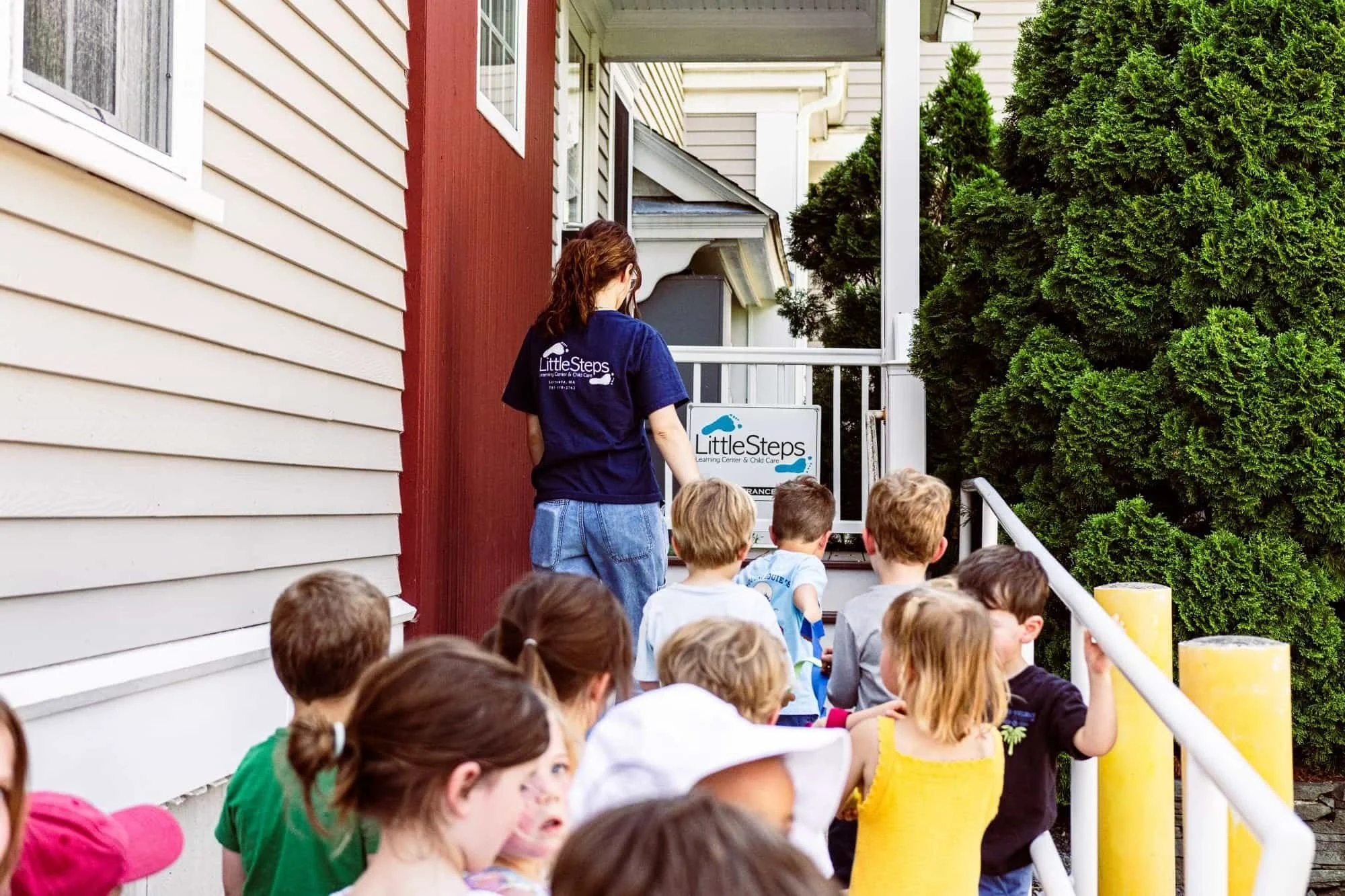 A group of children) waiting in line outside a building, while a woman in an uplifting shirt guides them. The building has beige and red siding, and there are large green bushes nearby. The scene suggests an outdoor group activity or educational trip.
