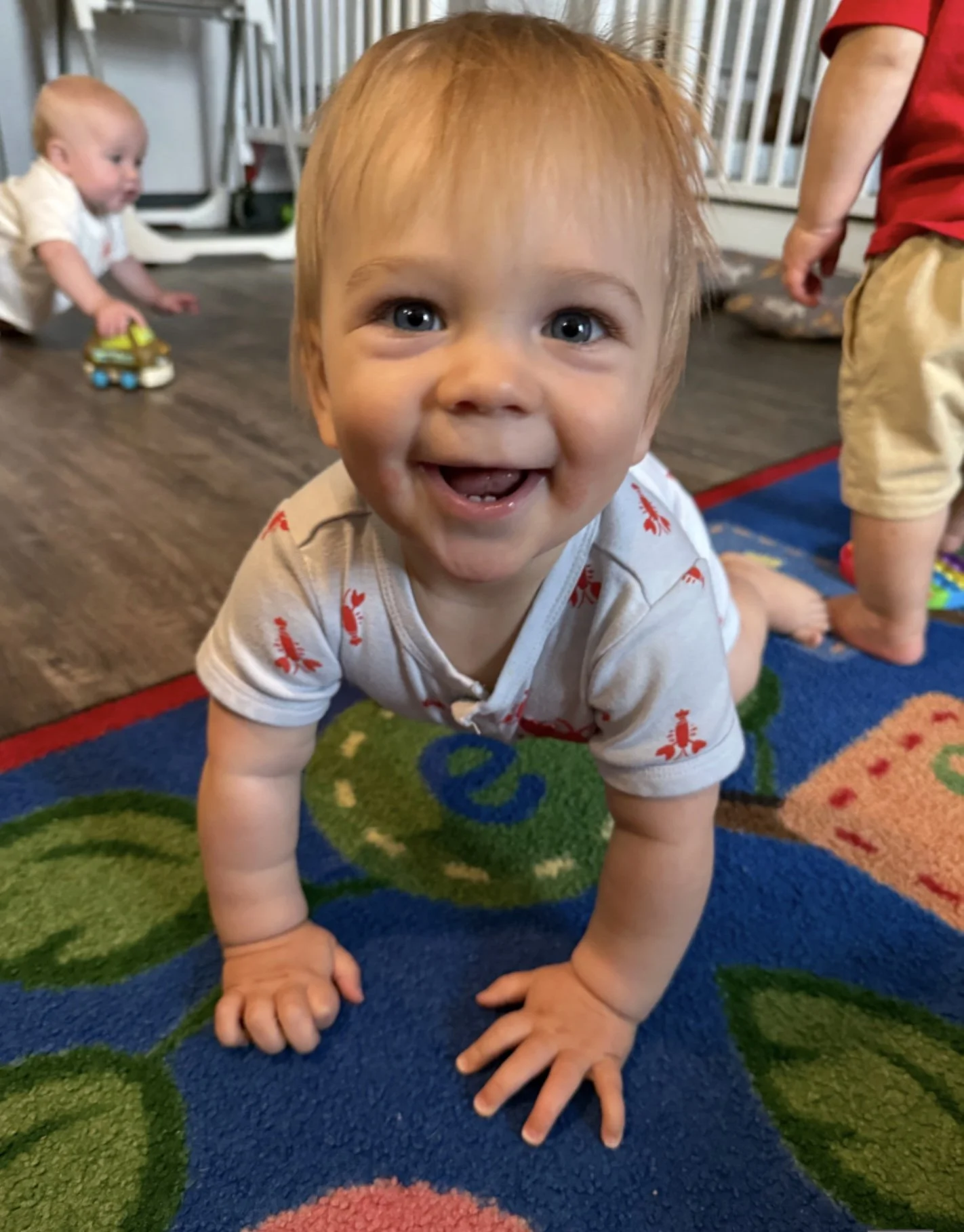 A smiling toddler with red hair crawling on a colorful rug, with other children playing in the background.