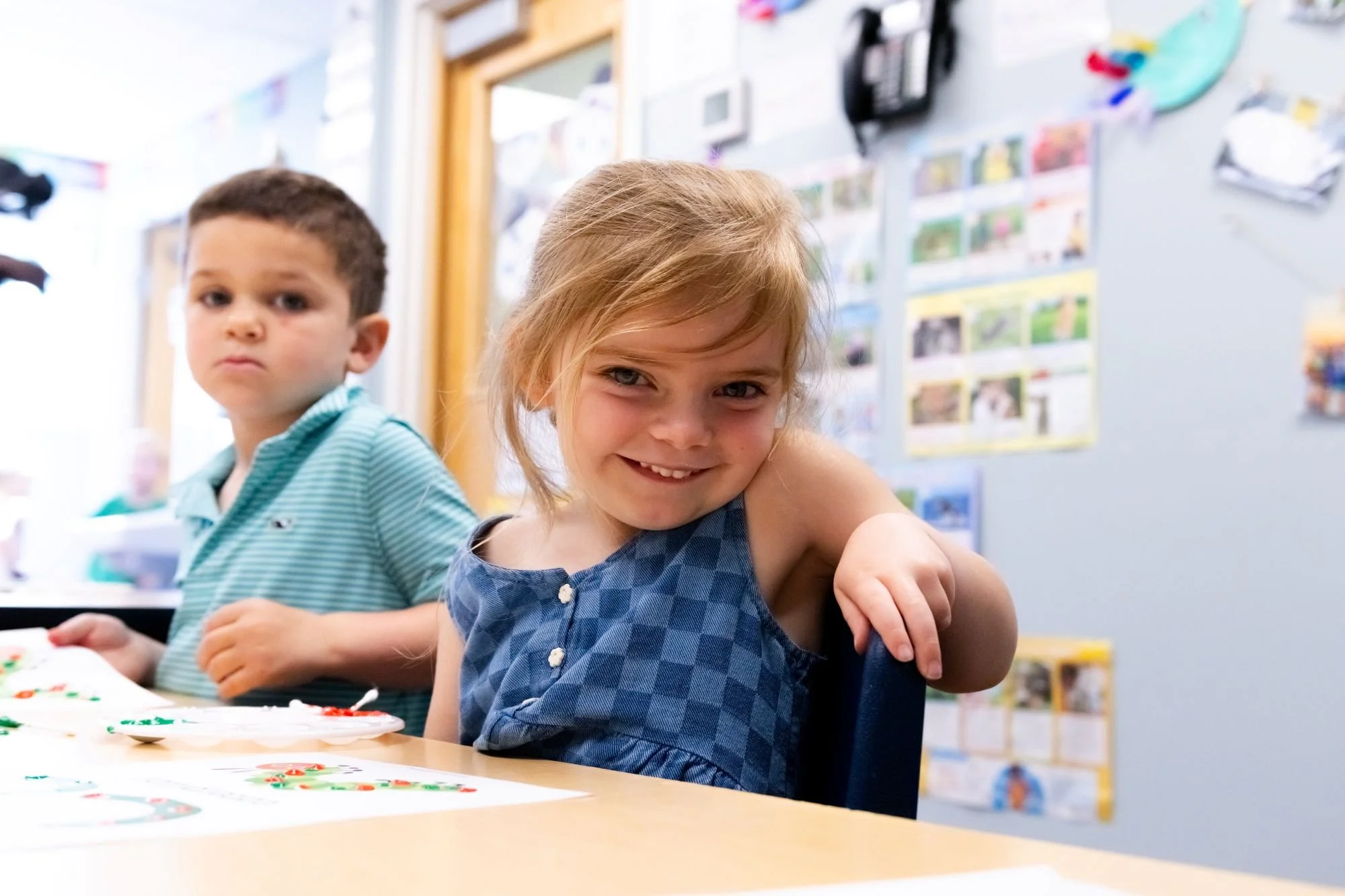 Young girl with red hair smiling and sitting at a table with her arm around her friend, a young boy, in a classroom decorated with colorful pictures and posters on the walls.