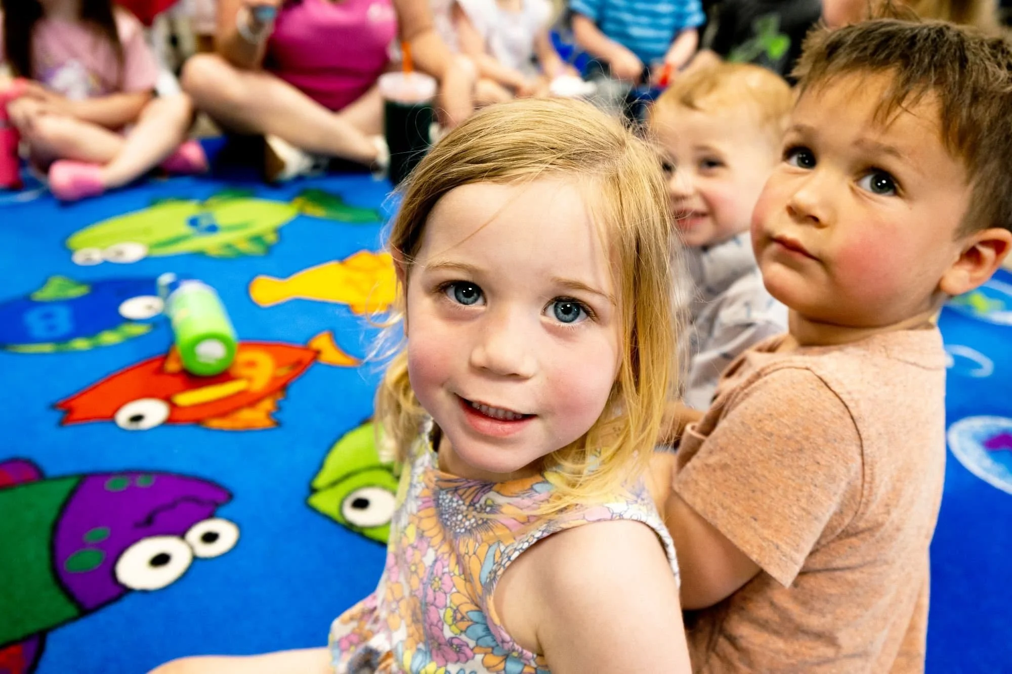 Children sitting on a colorful, cartoon-themed carpet in a classroom or play area.