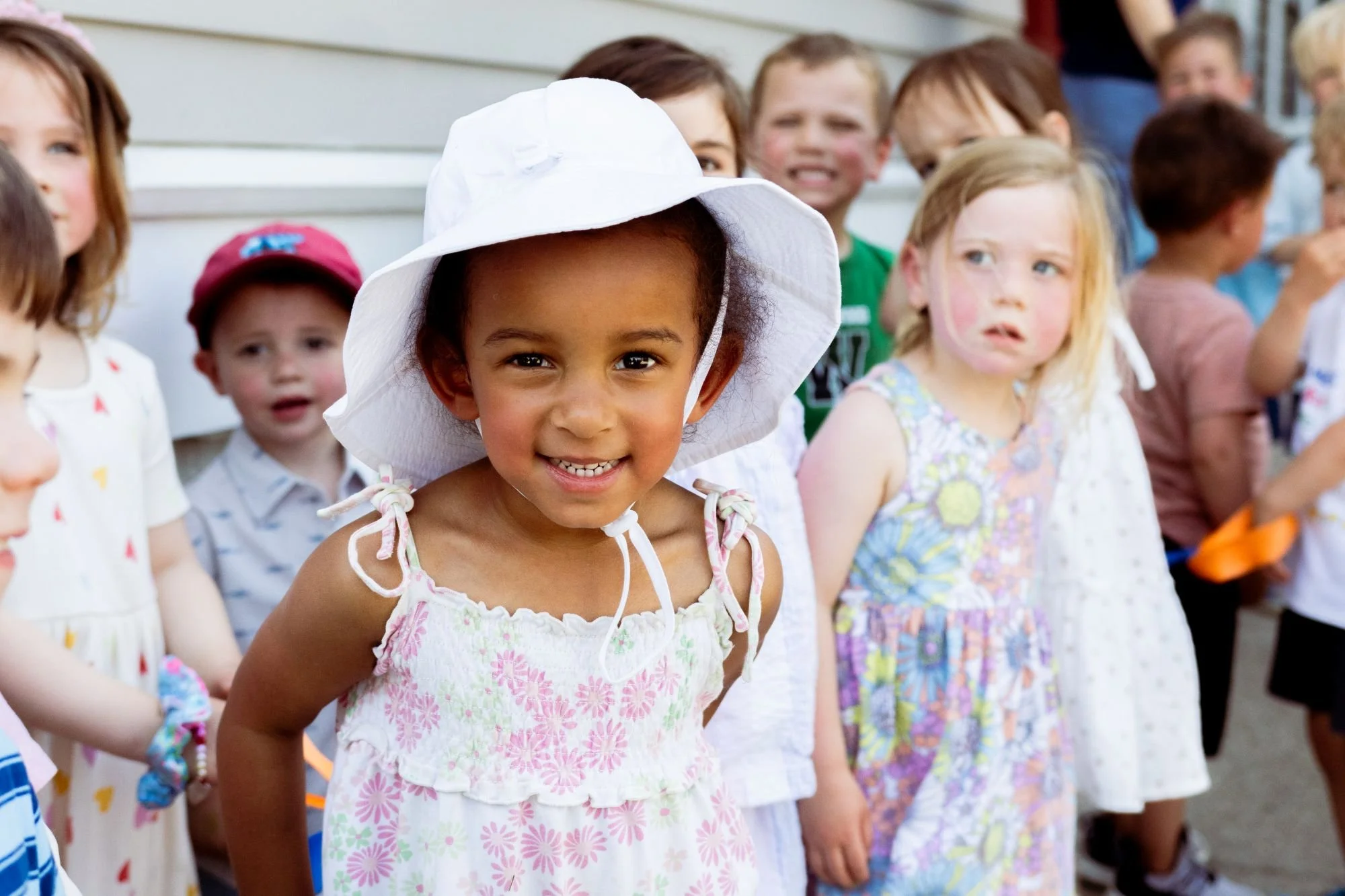 A group of children standing outdoors, smiling and looking at the camera, with a girl in a white sun hat and pink floral dress in the foreground.